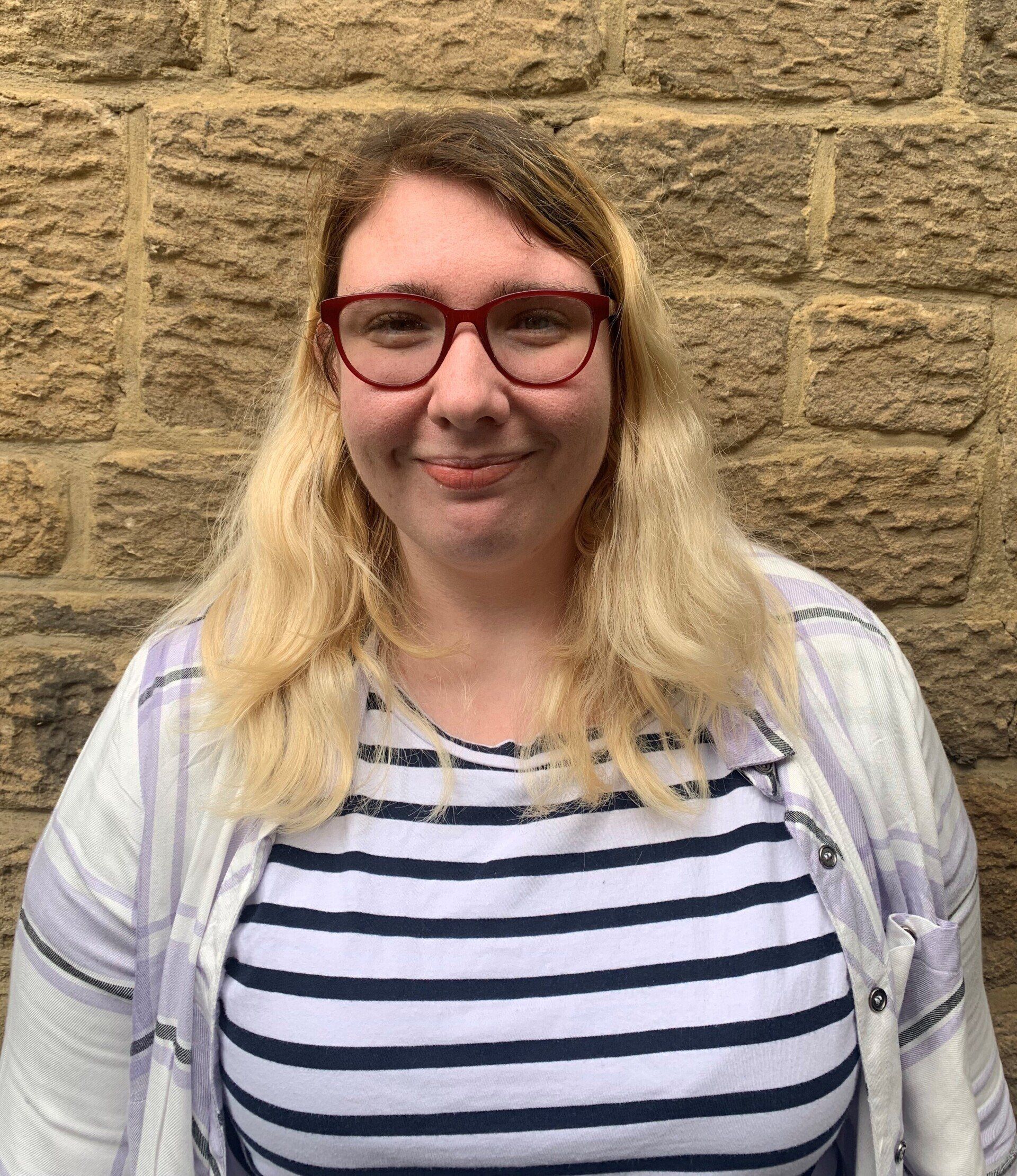 A photo of Victoria, who has long, slightly curly blond hair. Victoria is wearing glasses and has a striped top on.