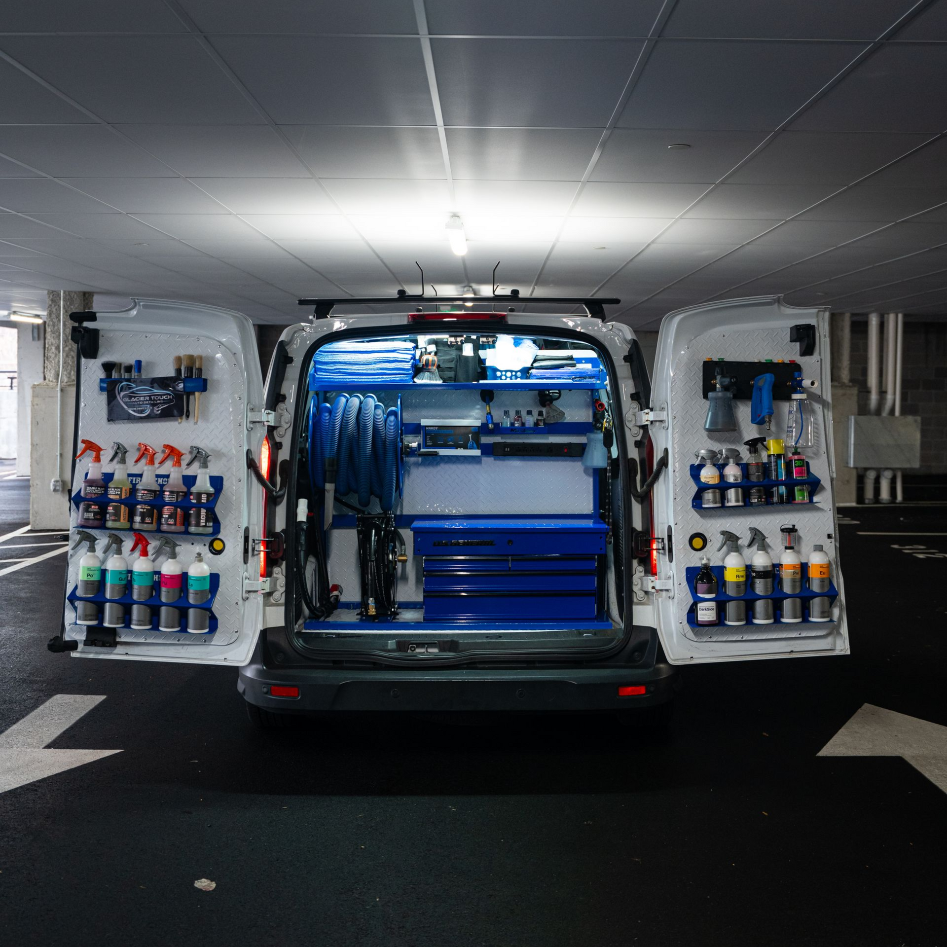 Open cargo van under garage lights, rear doors open with organized cleaning supplies and tools inside.