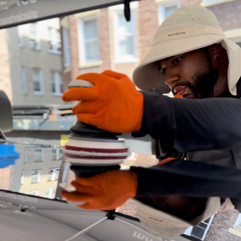 Person in a beige hat and orange gloves carefully cleaning a car windshield outdoors.