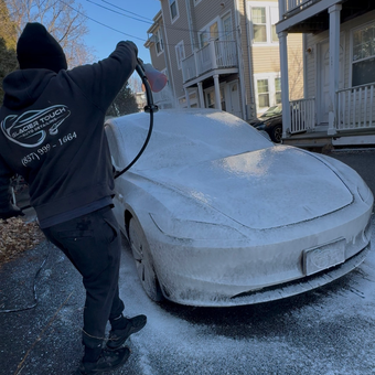 Person spraying snow off a snow-covered car on a residential street