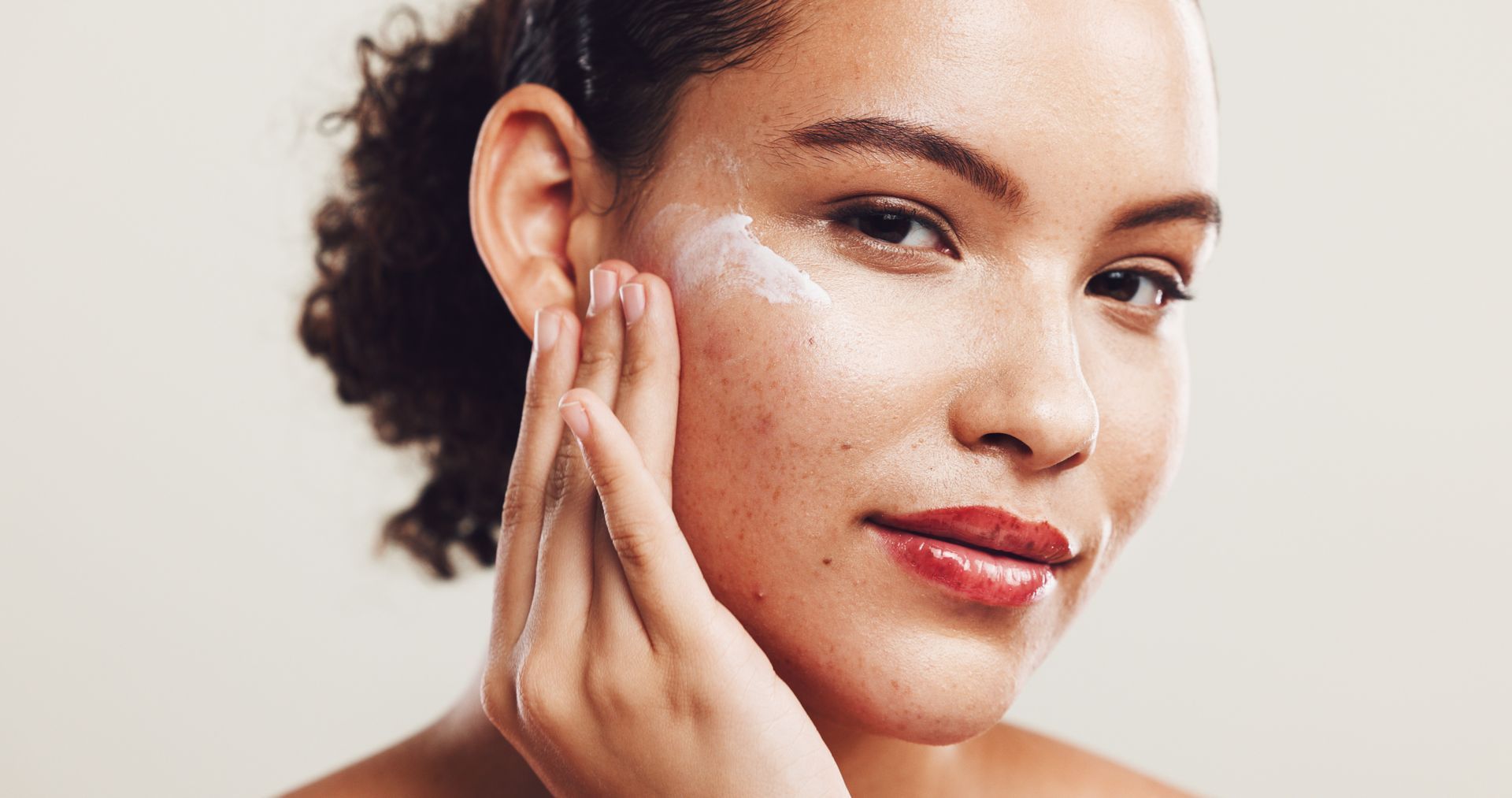 A person gently applying a white cream to their cheek against a neutral background.