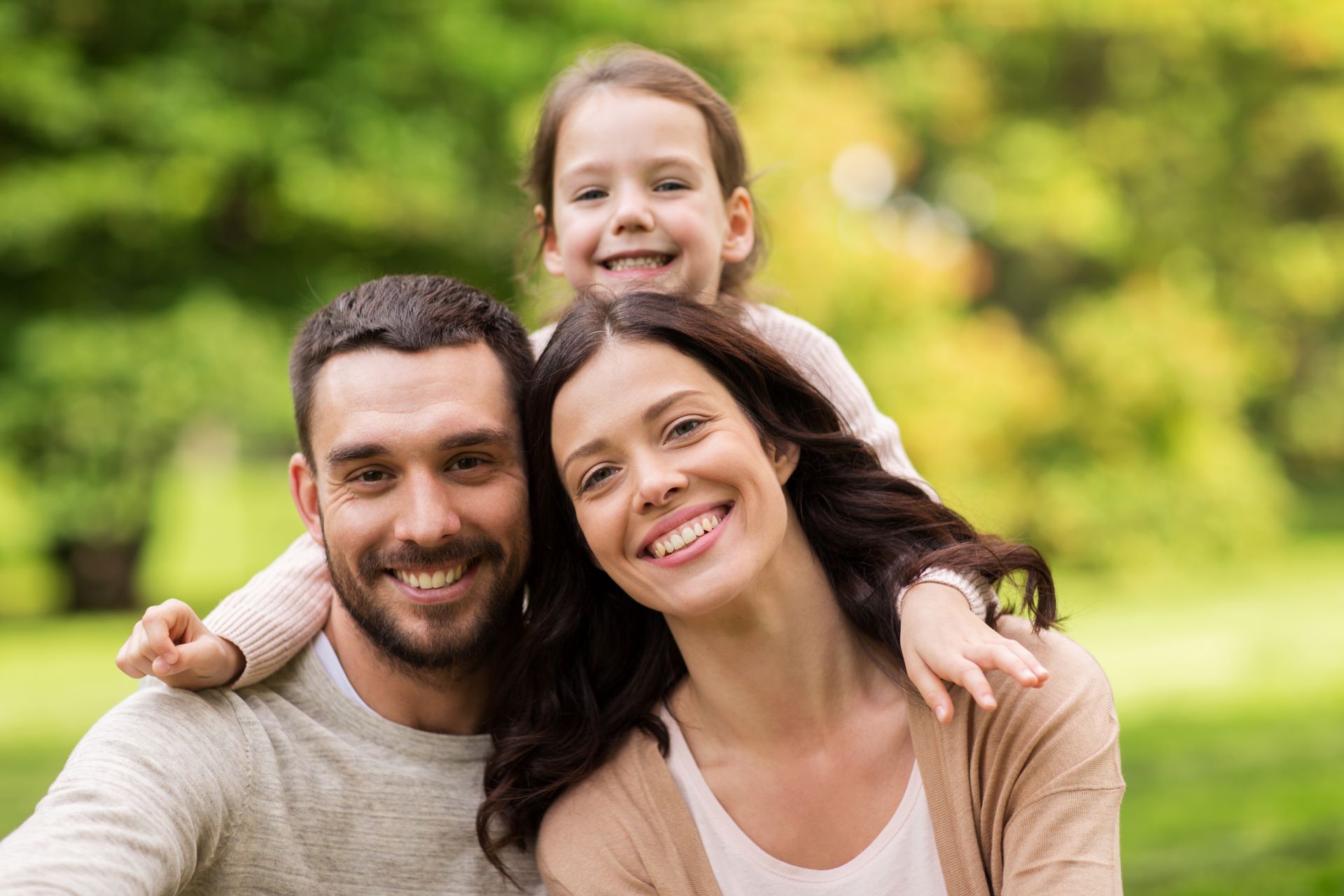 Smiling family of three poses outdoors: a child leans on parents, all looking at the camera. Green background.