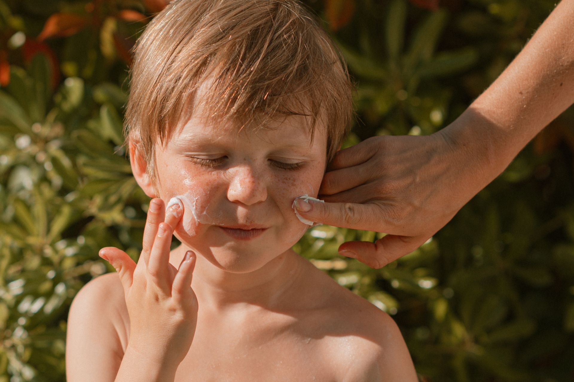 A person applying sunscreen to a child's face outdoors, near green foliage. 
