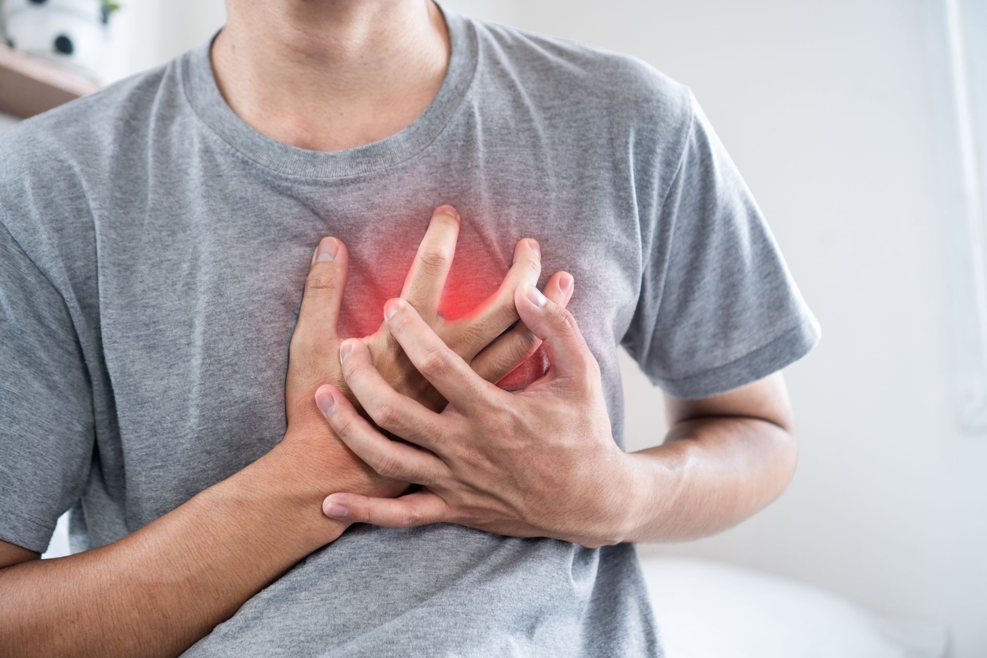 A person in a grey t-shirt holding their chest with both hands, where a red glow highlights the area of chest pain.