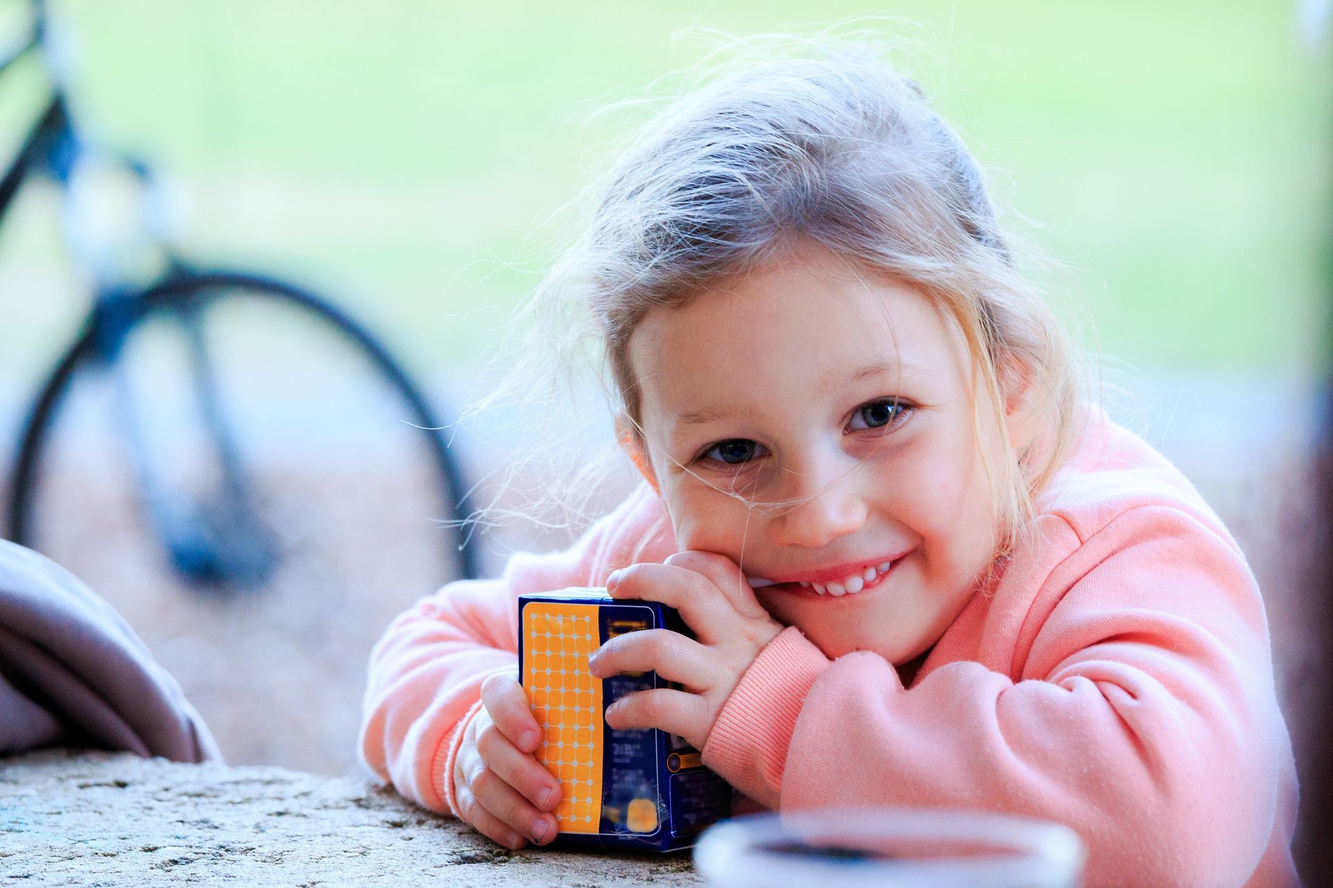 A person wearing a pink sweater smiles while holding a small, orange-and-blue juice box at a table, with a bicycle behind.