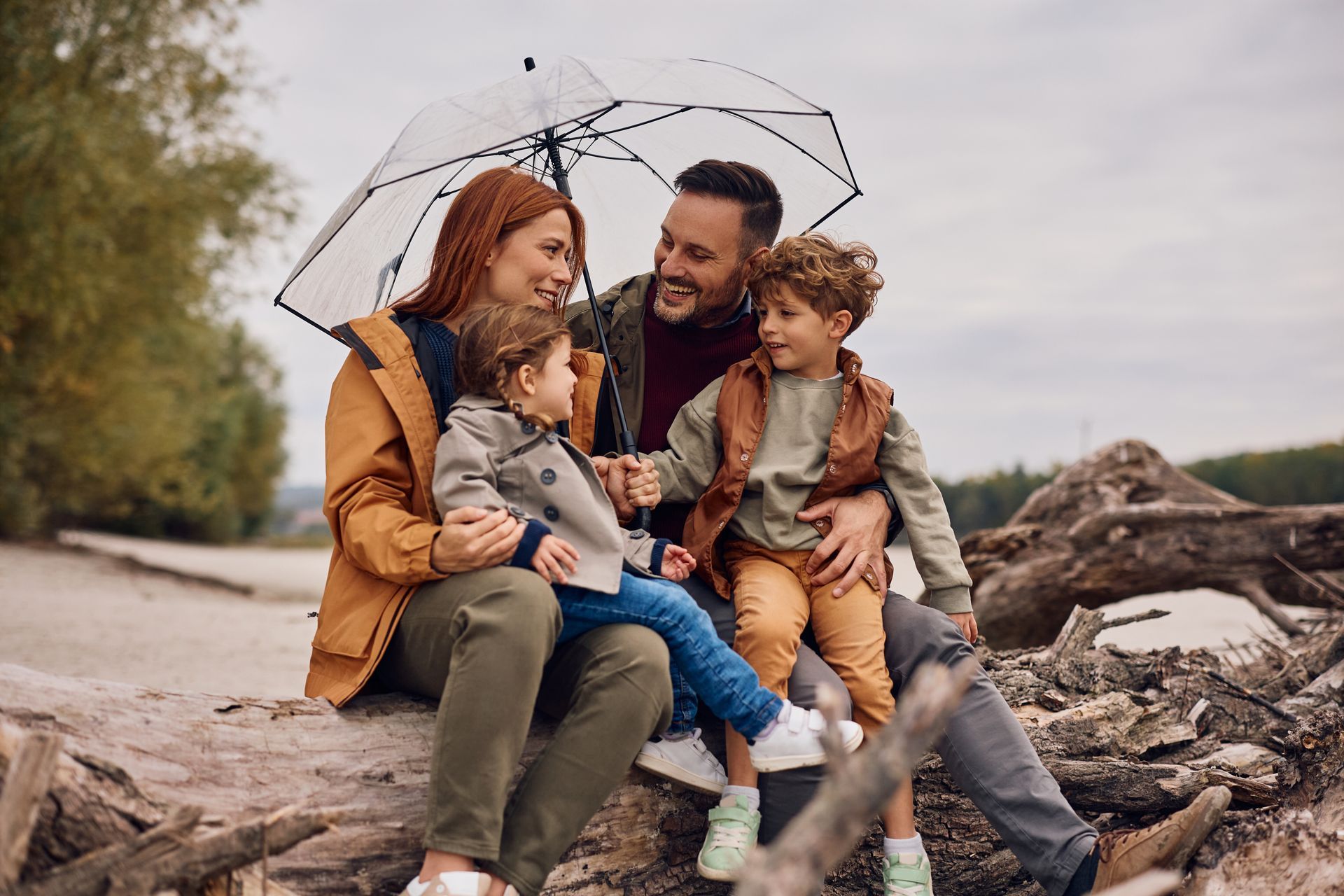 A family sitting on a large log outdoors under a clear umbrella on an overcast day.