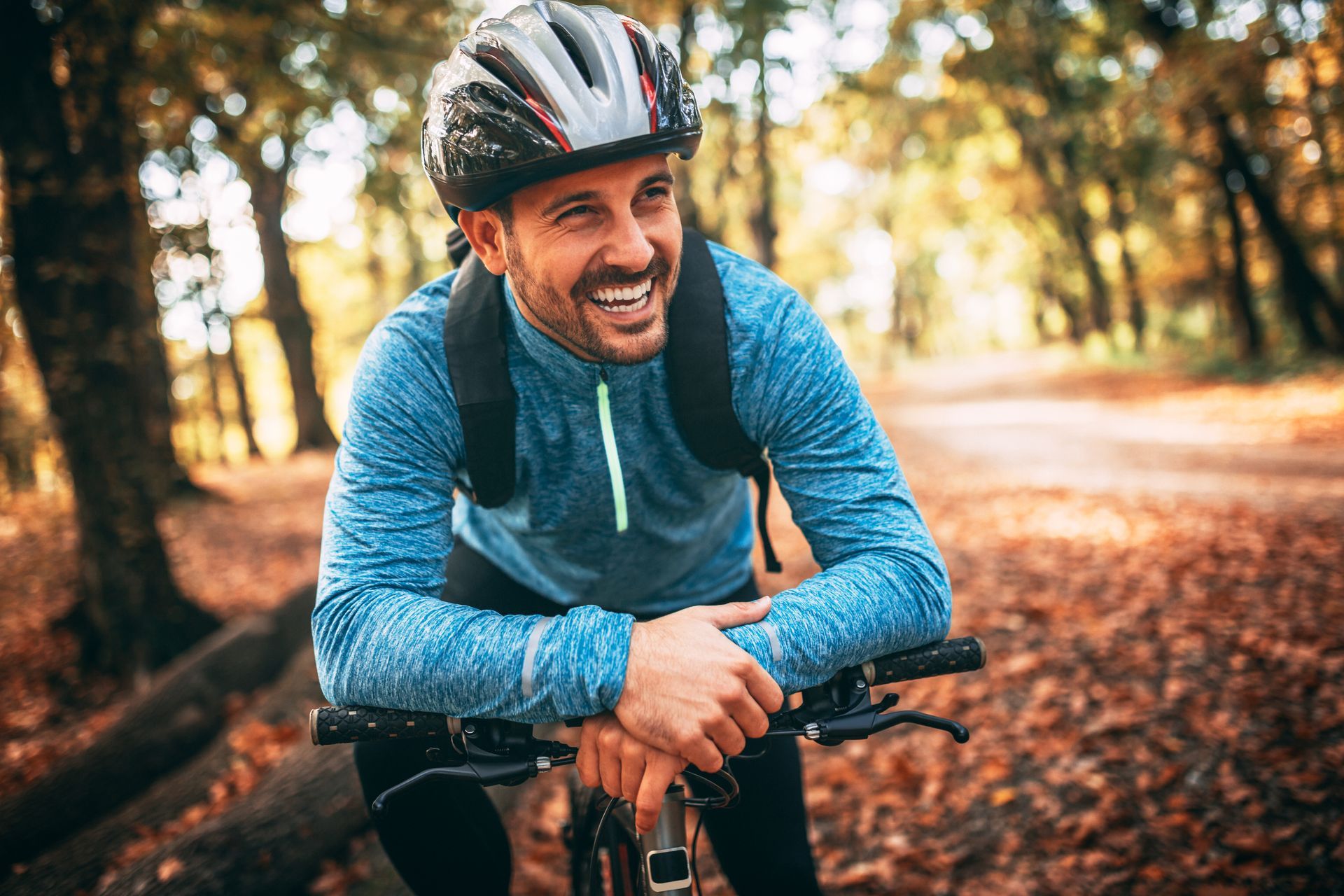 Man in bike helmet smiling, resting on handlebars in a colorful autumn forest.