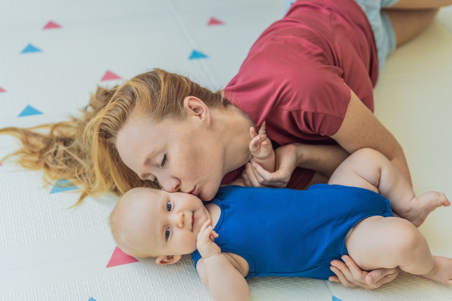 A person with long hair lies on a patterned floor, tenderly kissing a baby wearing a blue bodysuit.