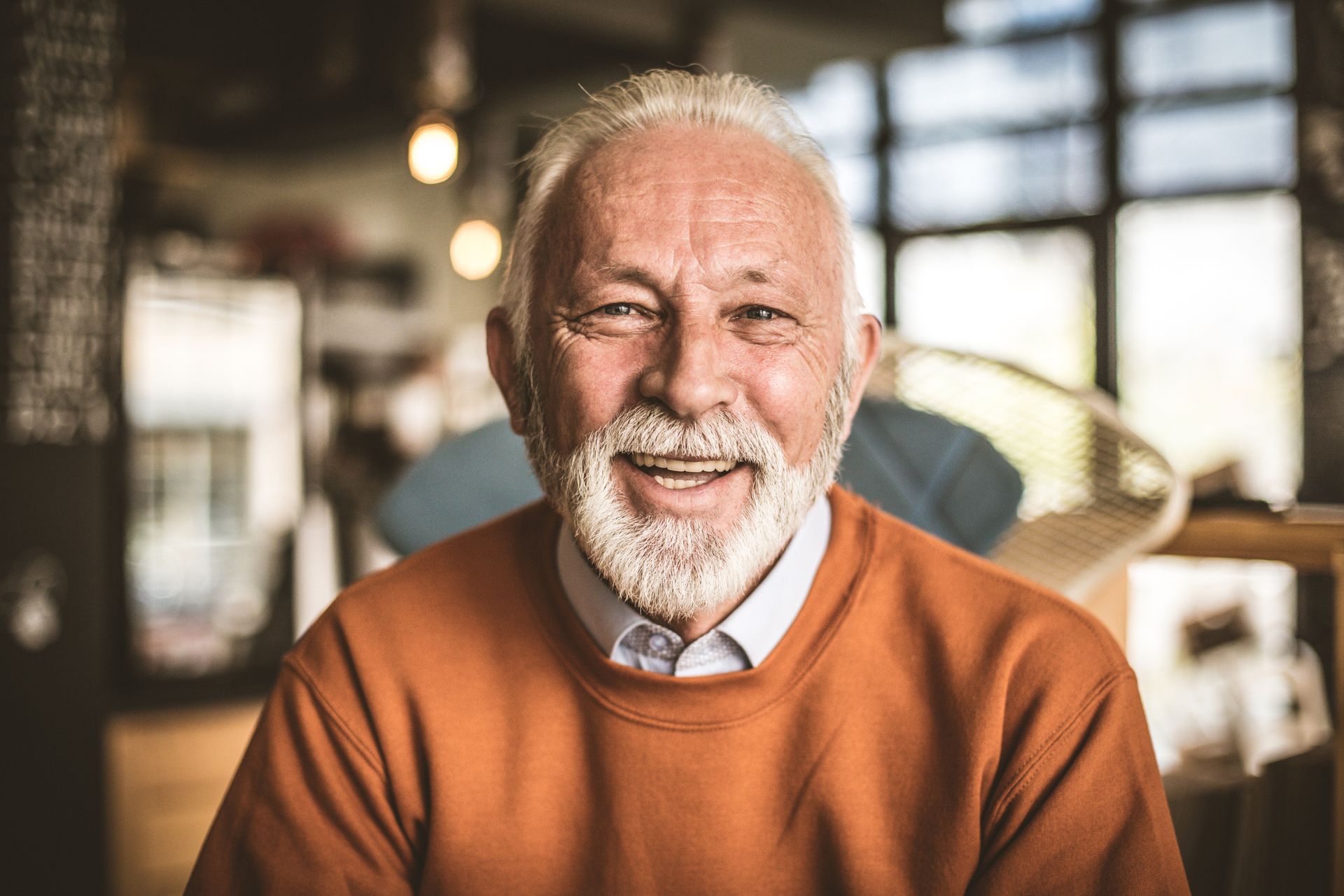 Smiling older adult with a white beard and orange sweater, indoors.