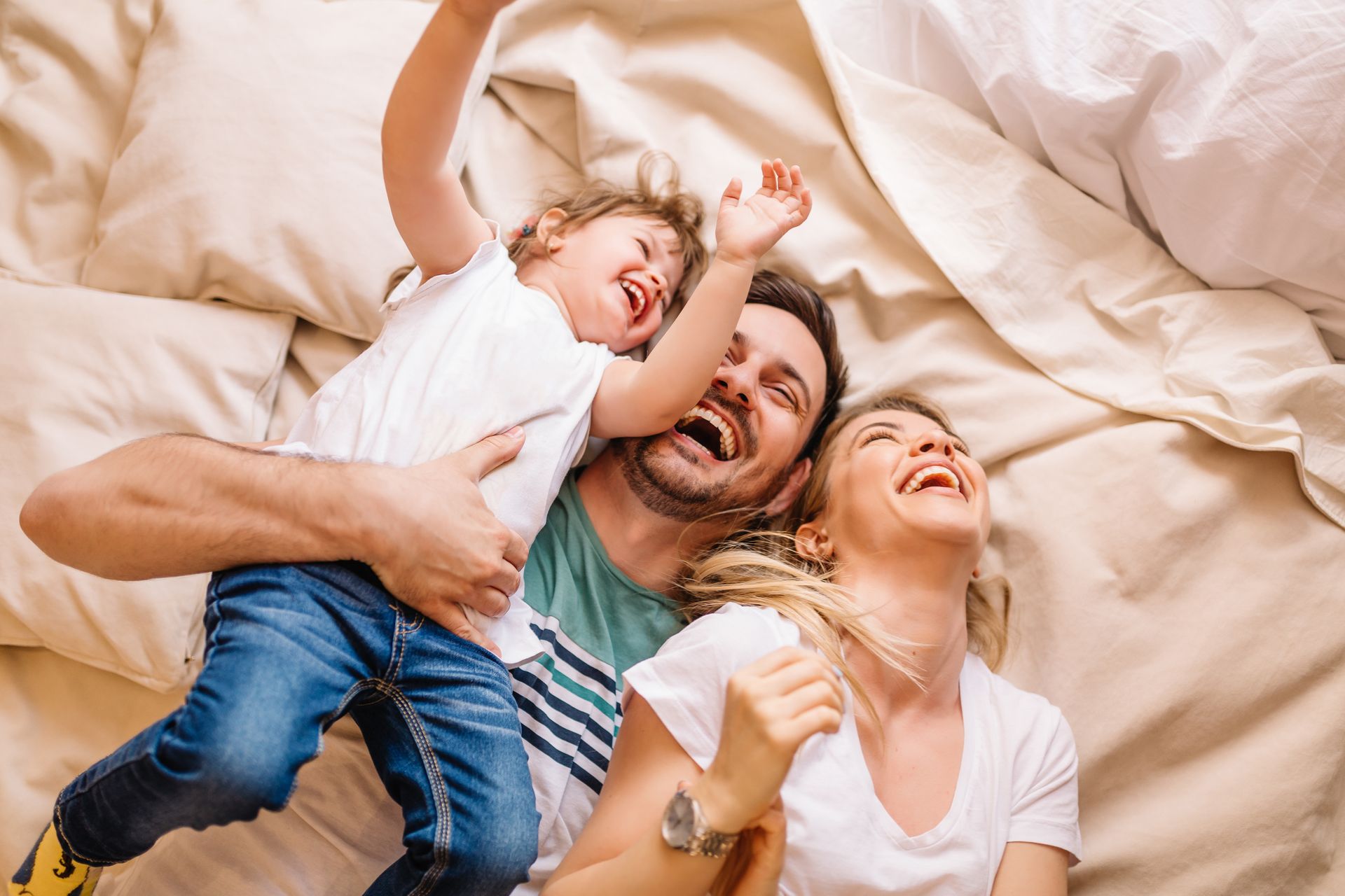 Family laughing on bed, child held by adult. Light bedding, joyful expressions.