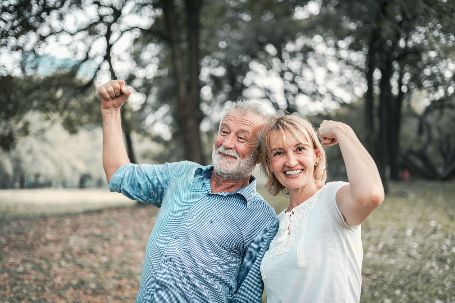 Smiling couple flexing their arms outdoors, trees in background.