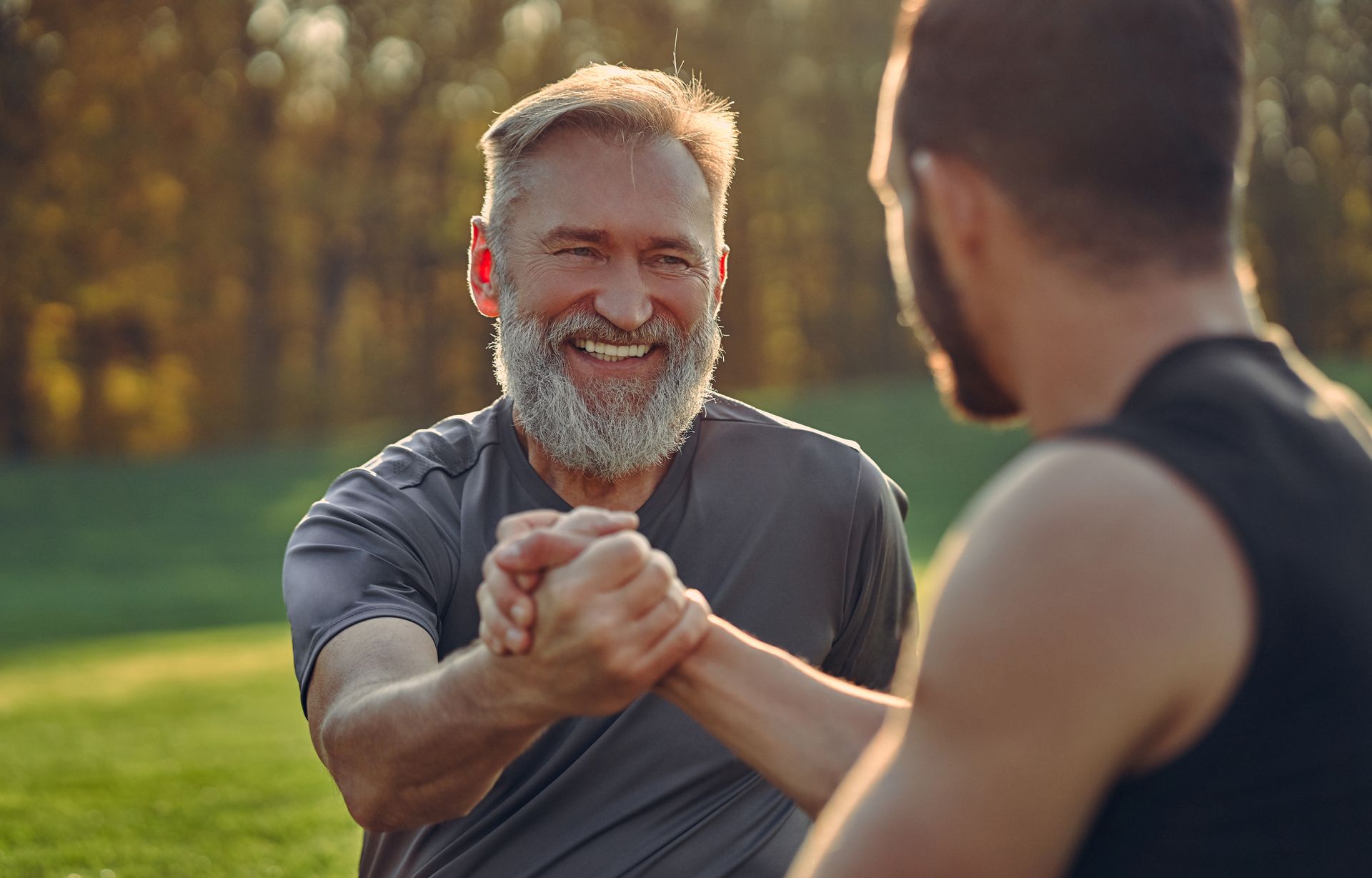 Two people shake hands outdoors on a grassy field, smiling at each other during a workout in the sunlight.