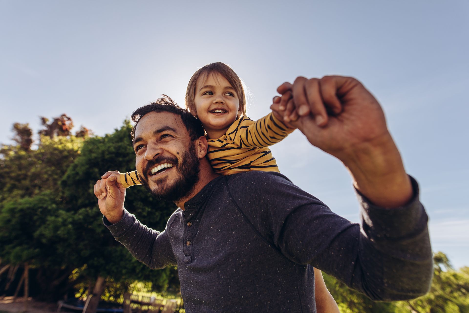 Man smiling, carrying a child on his shoulders outdoors.