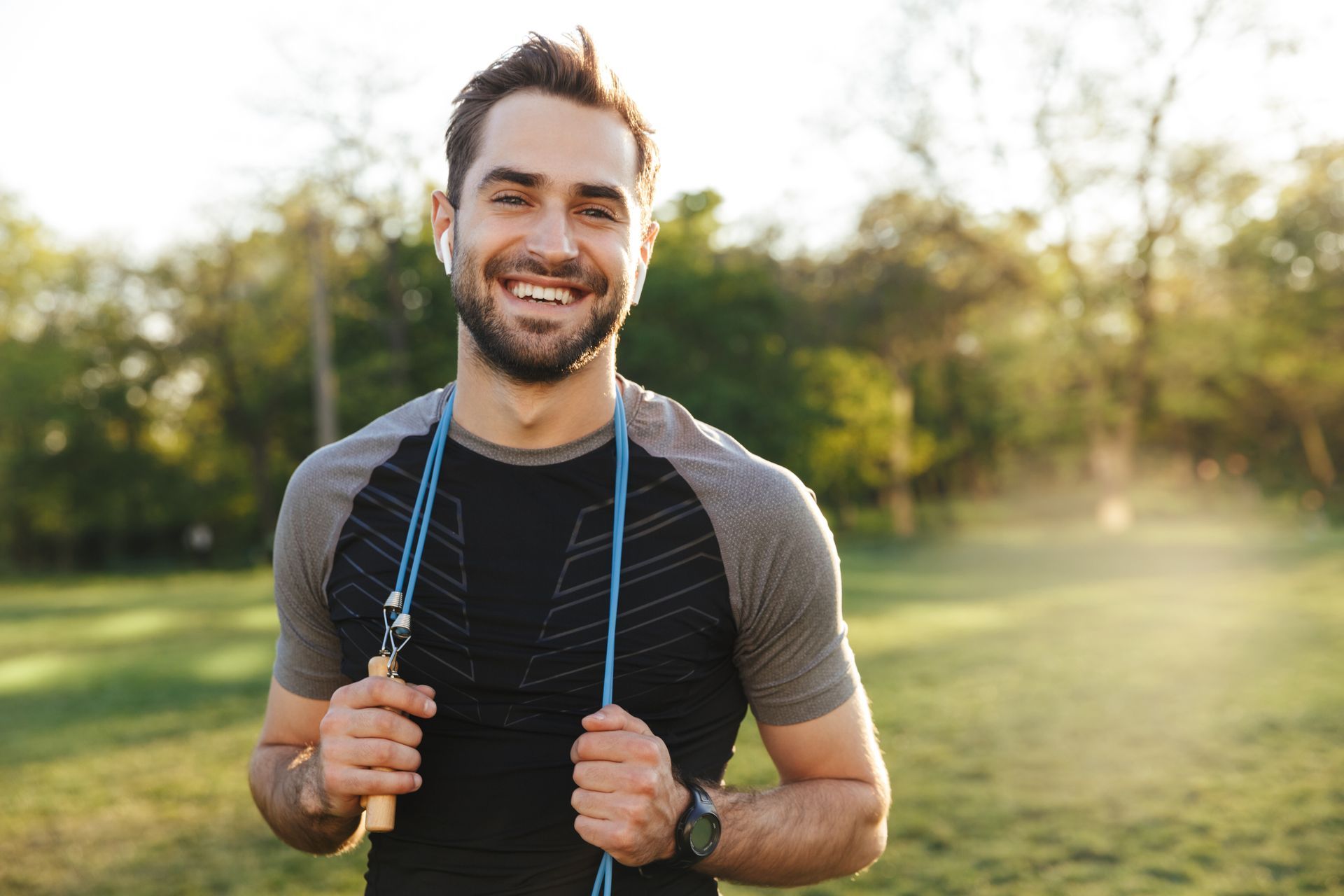 A smiling athlete in athletic wear holding a jump rope around their neck, standing in a sunlit park.