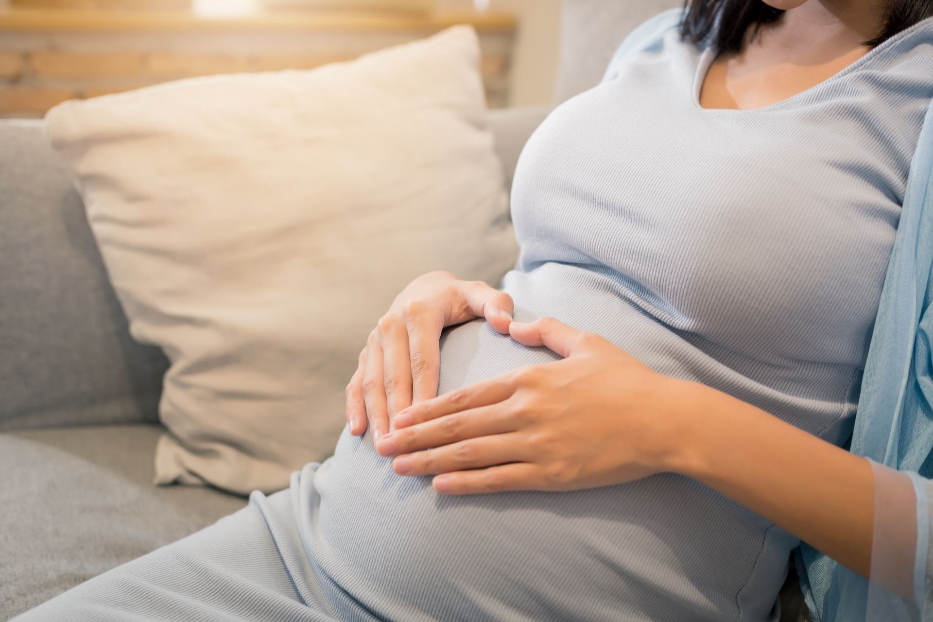 Pregnant person sitting, hands in heart shape on belly, wearing blue dress.