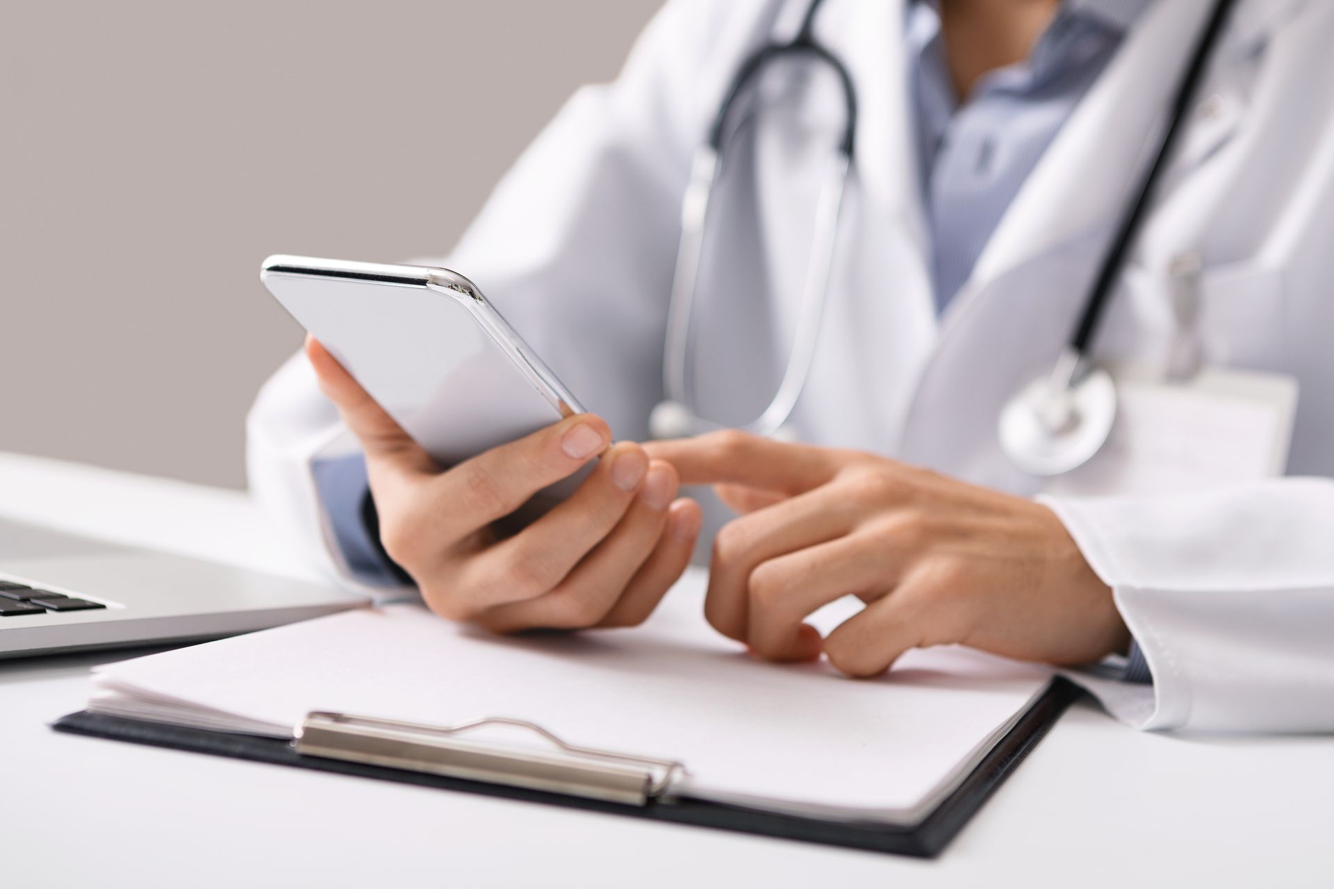 Doctor in white coat using a smartphone, stethoscope around neck, and clipboard on desk.