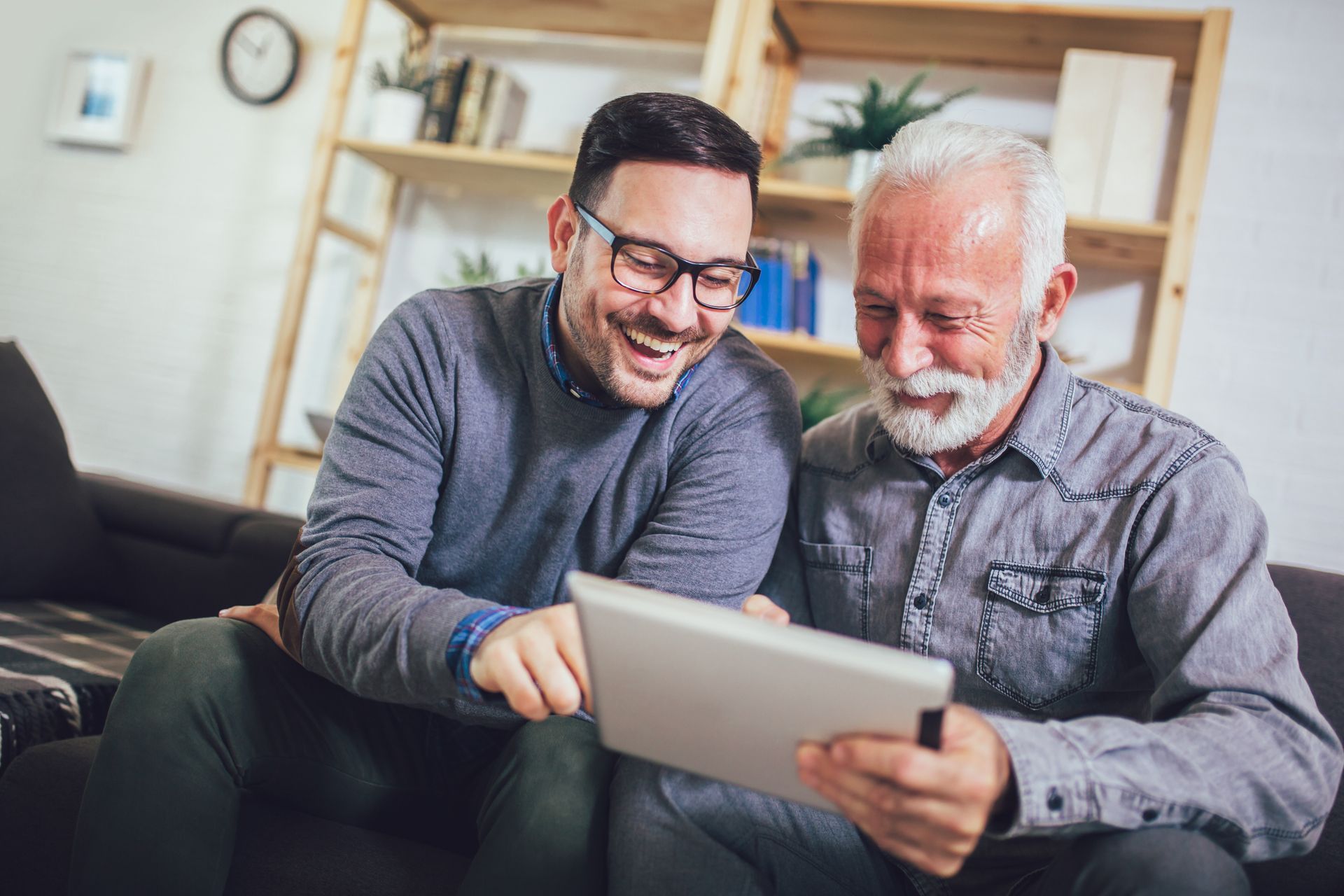 Man with glasses laughs while showing a tablet to an older man with white hair, both on a couch.