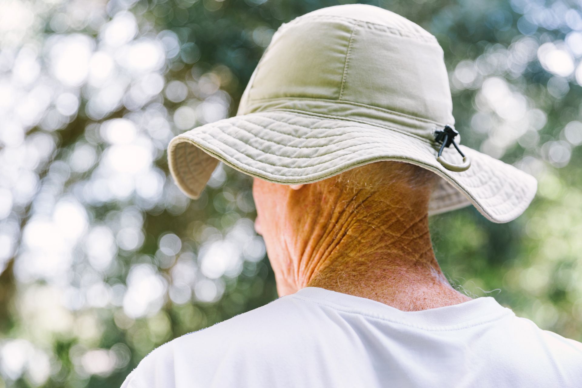 Person wearing a light-colored hat, seen from behind, with a wrinkled neck and blurred foliage background.