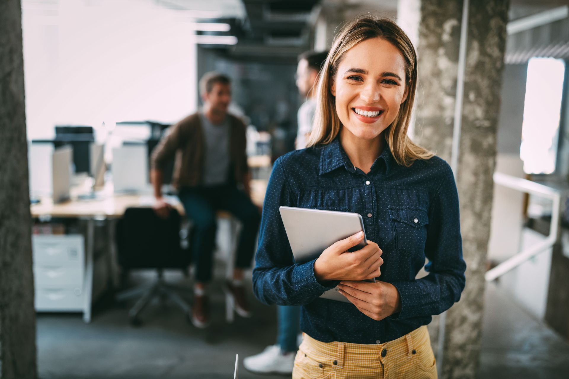 Woman smiling, holding tablet in modern office. Others blurred in background.