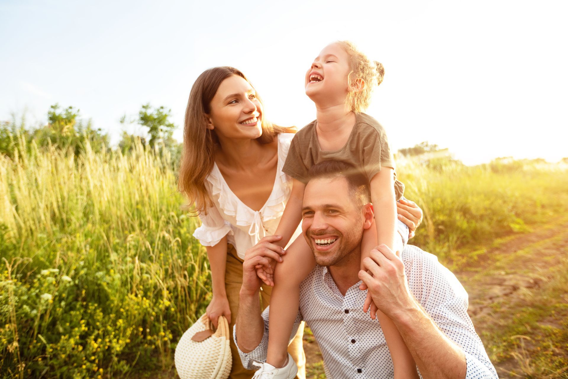 Family laughing outdoors: child on dad's shoulders, mom smiling, sunny field.