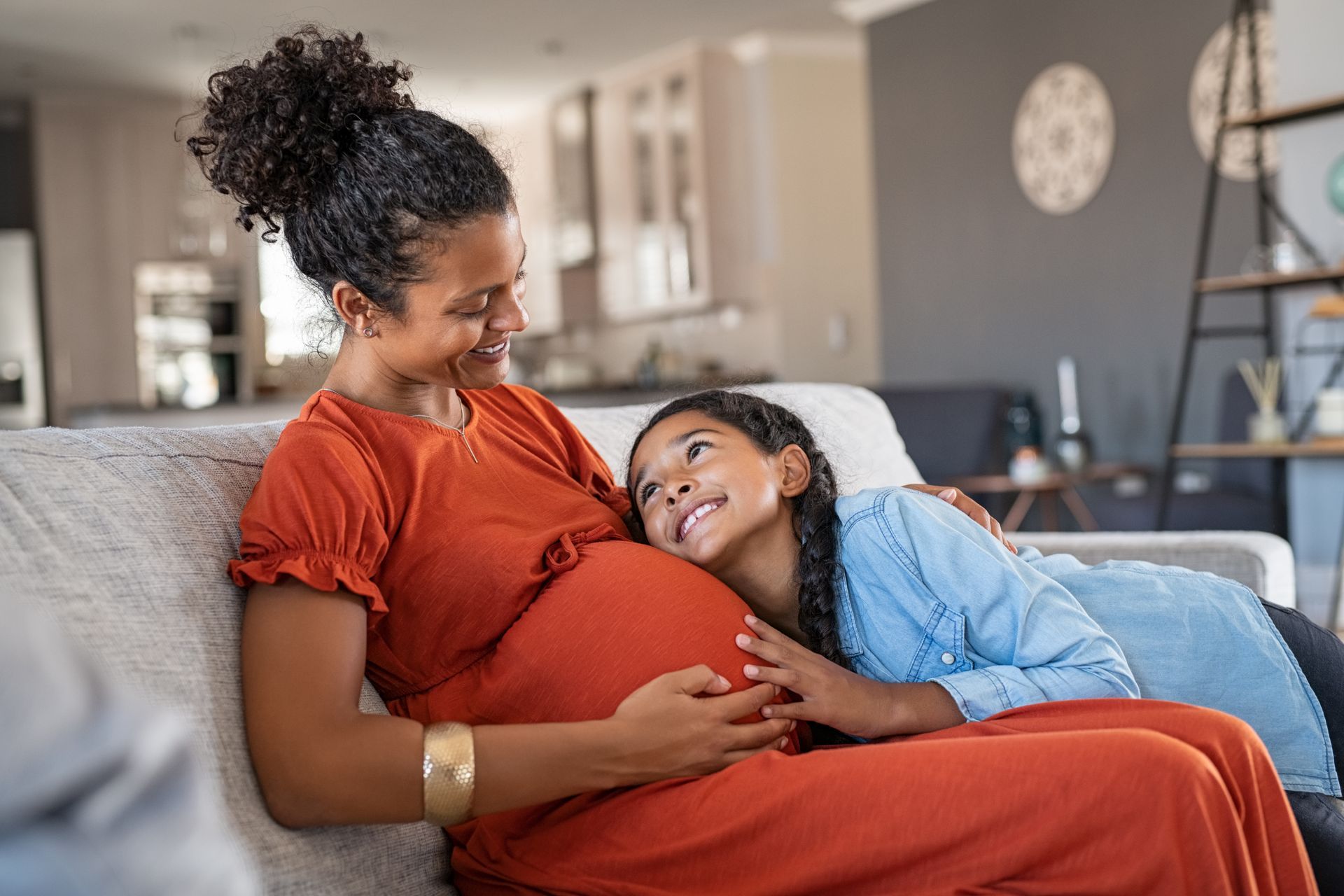 Pregnant person and child on couch, child listening to pregnant person's belly, smiling.