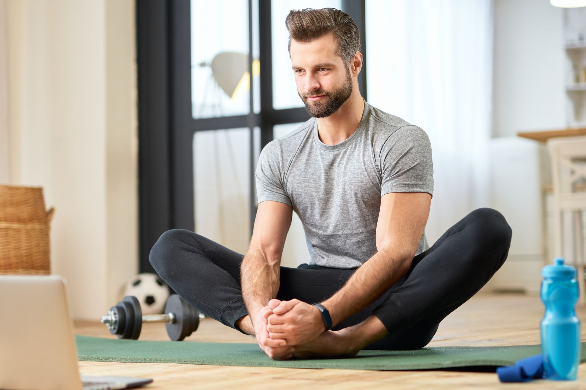 A person sitting on a green yoga mat, performing a butterfly stretch while looking at a laptop in a bright room.