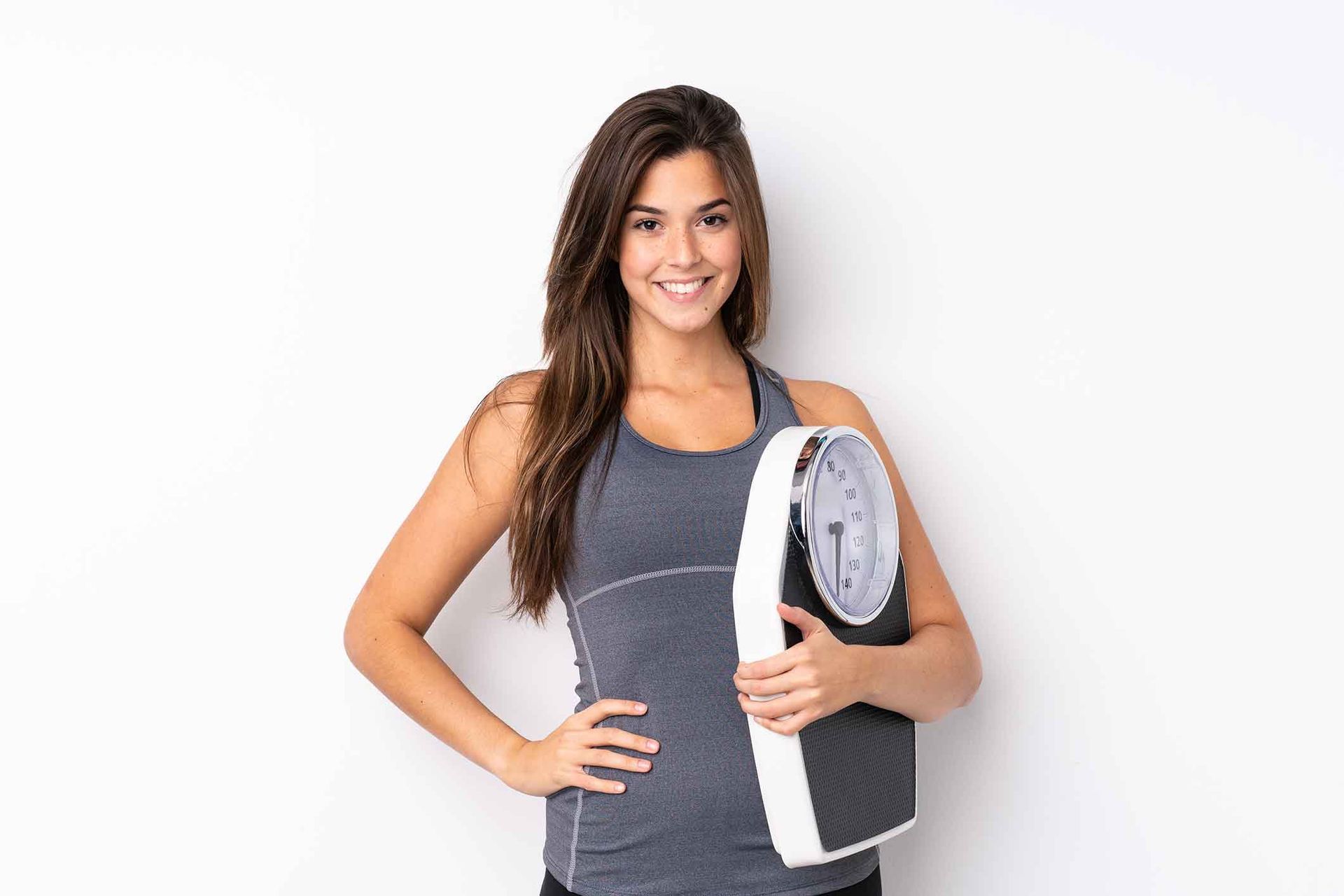 Woman in athletic wear smiling, holding a weight scale against a white background.