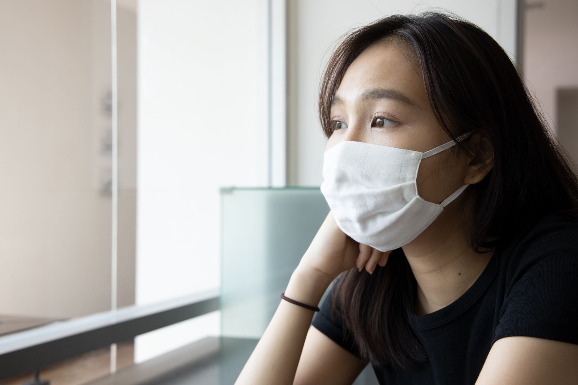 Woman wearing a white face mask looking out a window. She has dark hair and is wearing a black shirt.