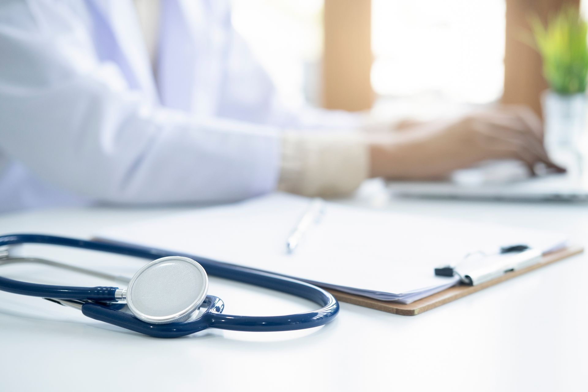 Stethoscope on desk in front of a doctor, notebook and pen, with the doctor working on a laptop in the background.