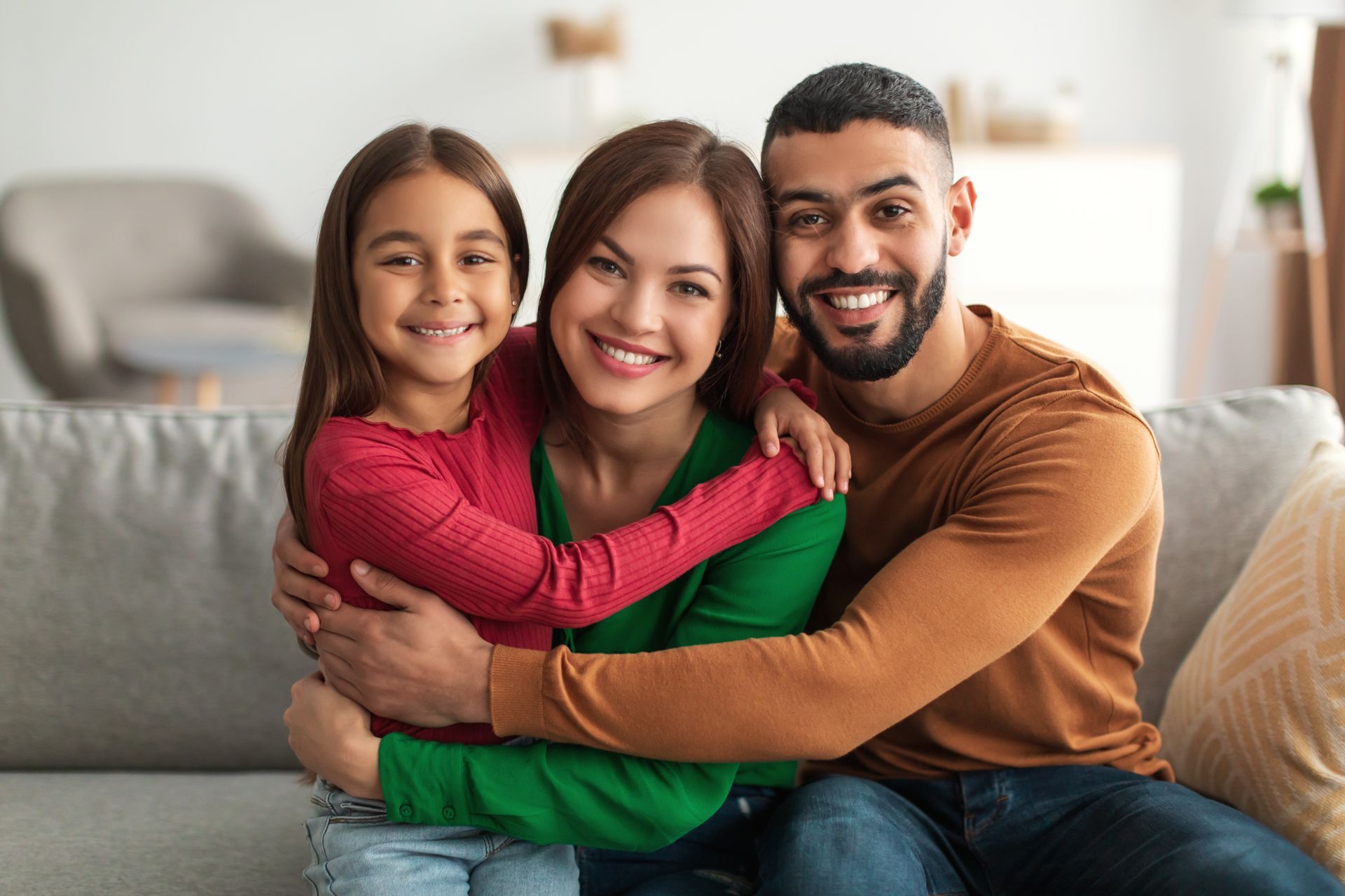 Family of three smiling, embracing on a couch indoors; mother, child, and father.