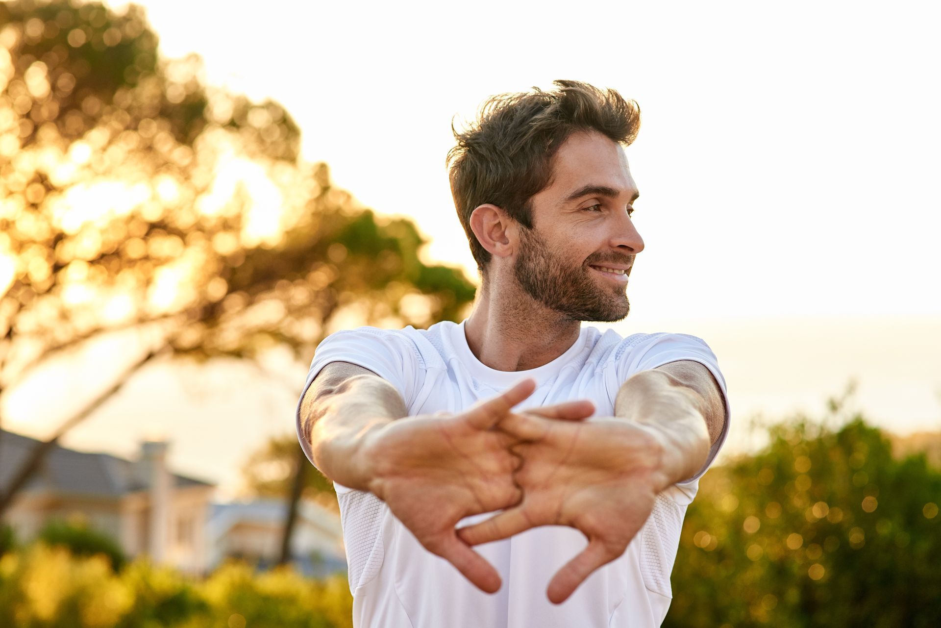 A person in a white t-shirt stretches their arms forward with hands clasped, smiling while looking away outdoors.