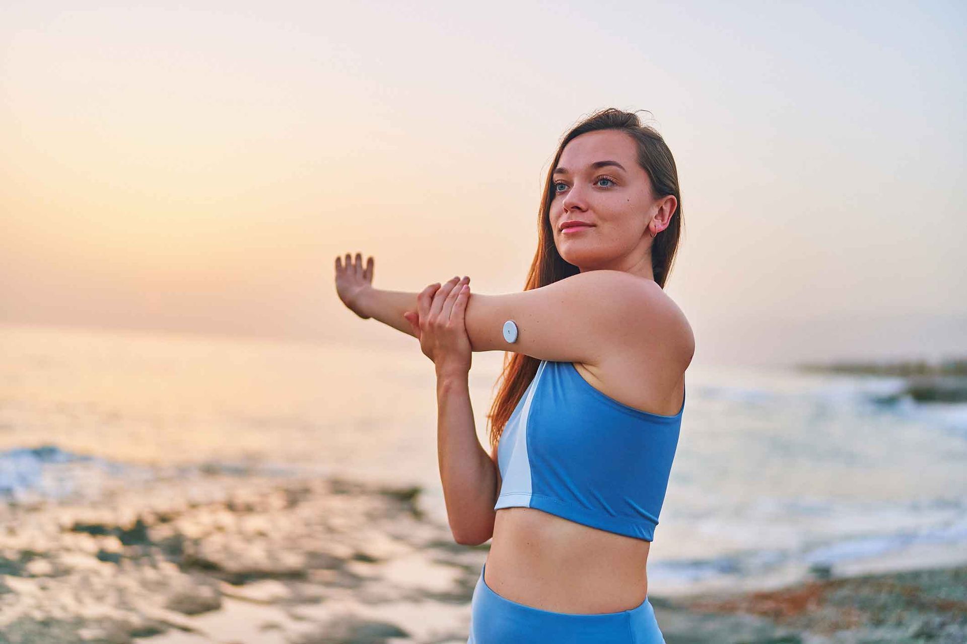 Woman in blue activewear stretches arm, ocean background, CGM on arm.