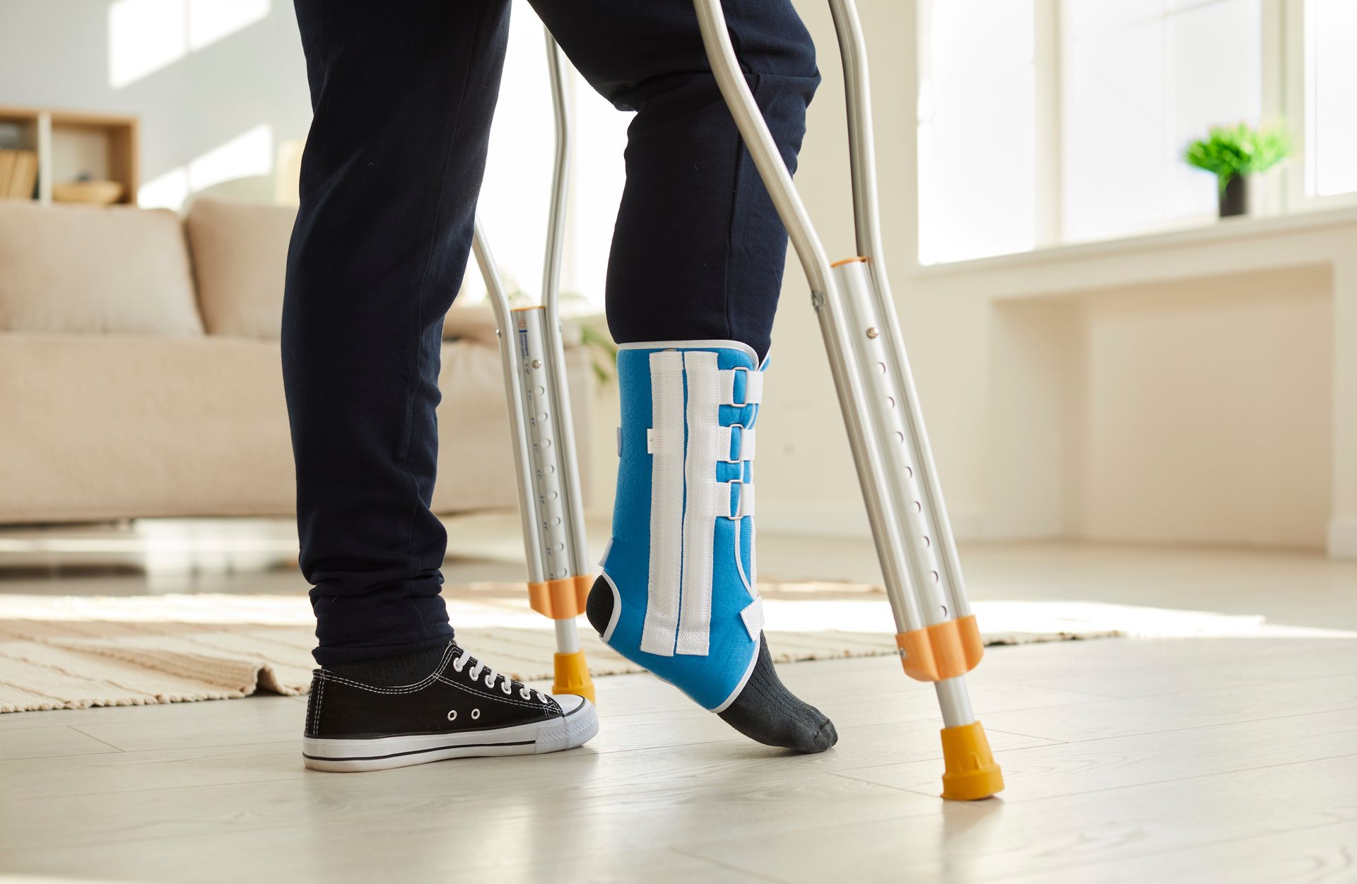 Person using crutches with foot in a blue and black medical boot indoors, near a couch and window.