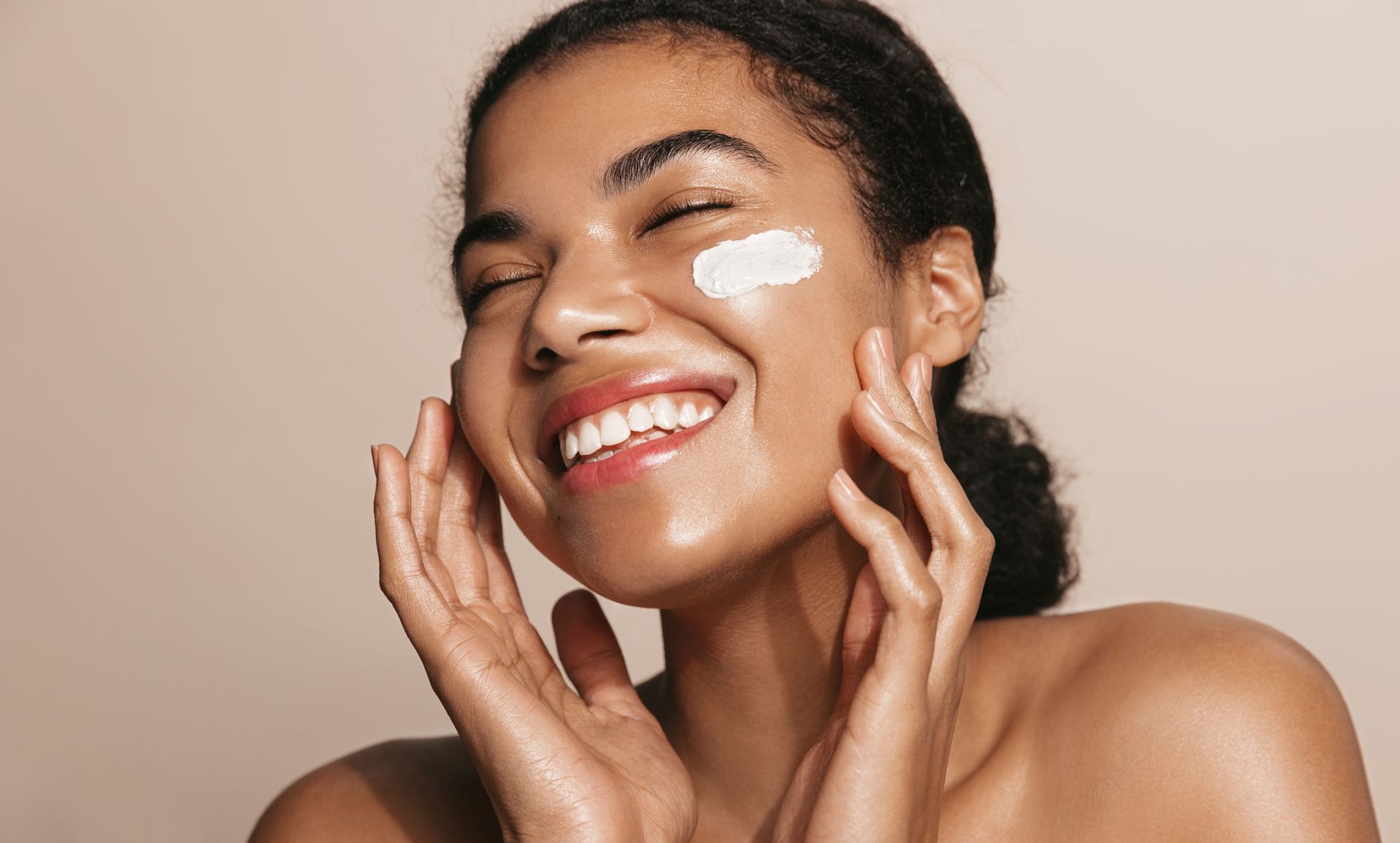 Woman applying cream to her face, smiling with eyes closed. Cream on cheek. Tan background.