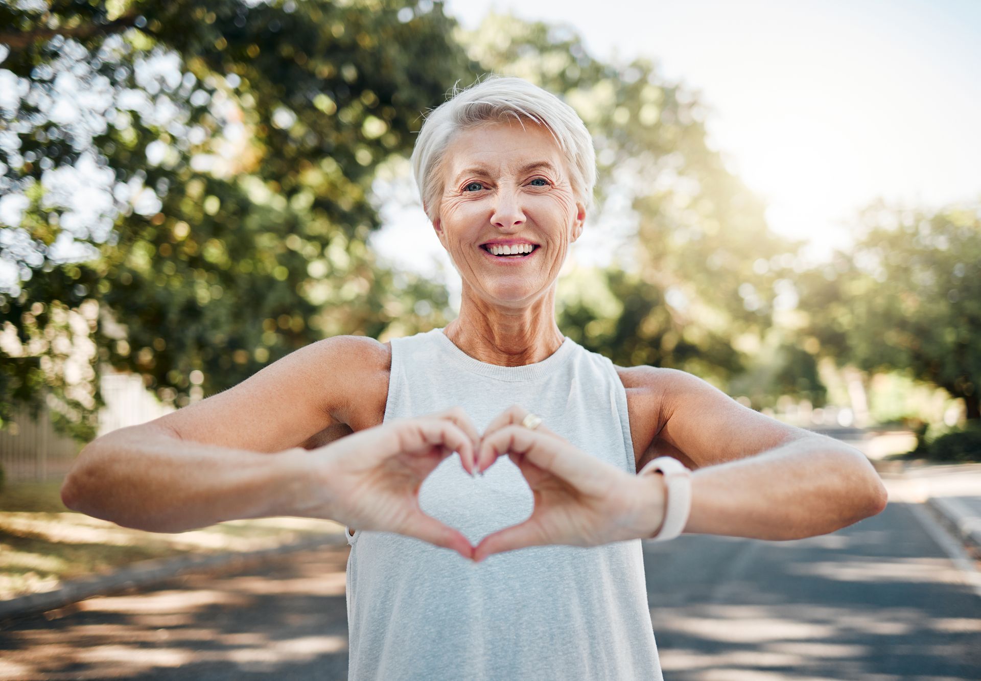 A person wearing a white tank top and a fitness tracker makes a heart shape with their hands outdoors in a park.