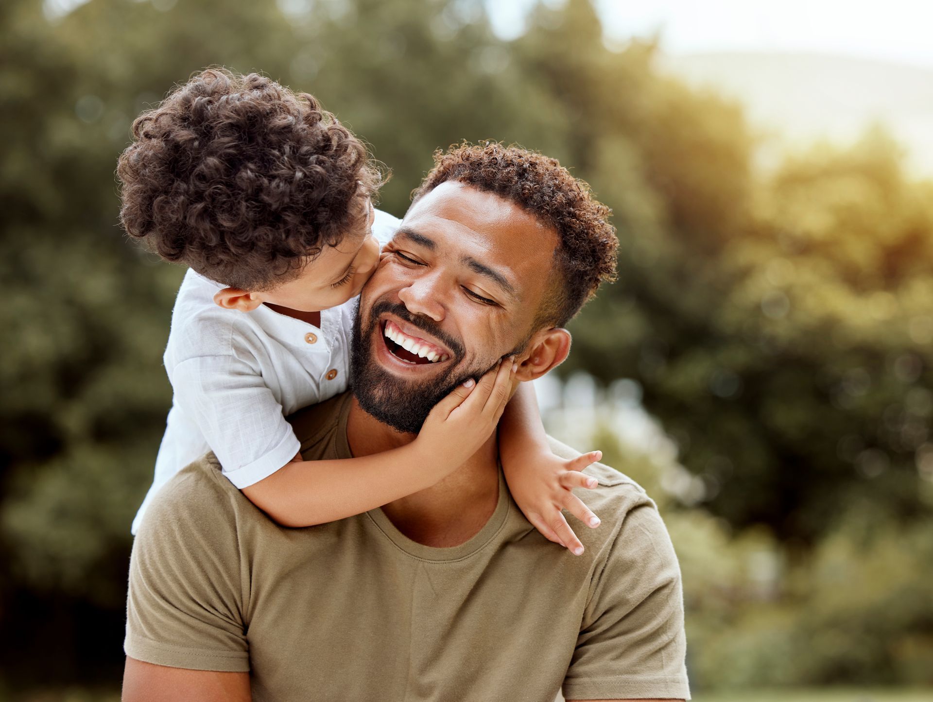 Boy giving a laughing man a kiss on the cheek outdoors, embraced in a hug.