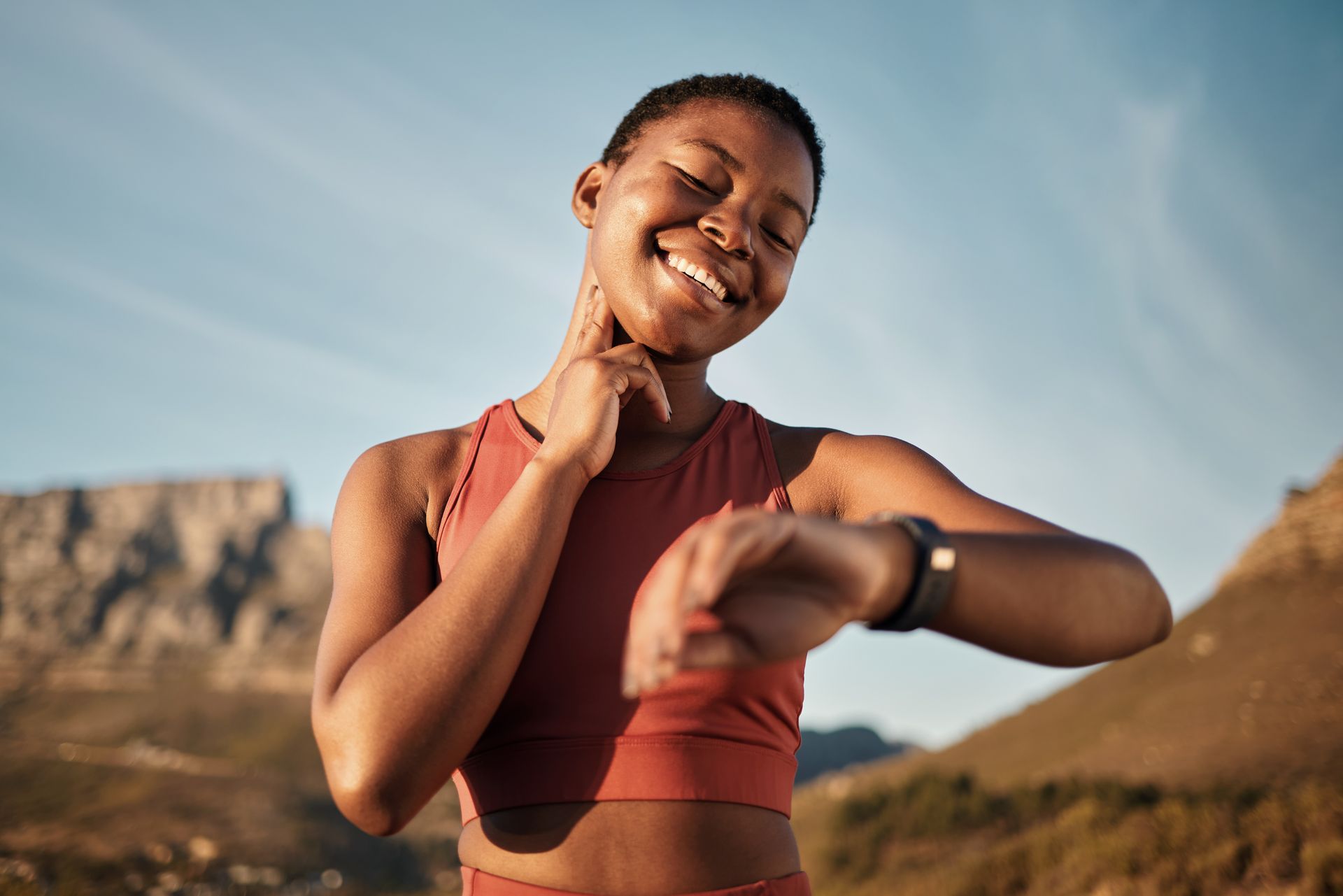 A person in a terracotta sports top checks their heart rate by holding their fingers to their neck, outdoors by mountains.