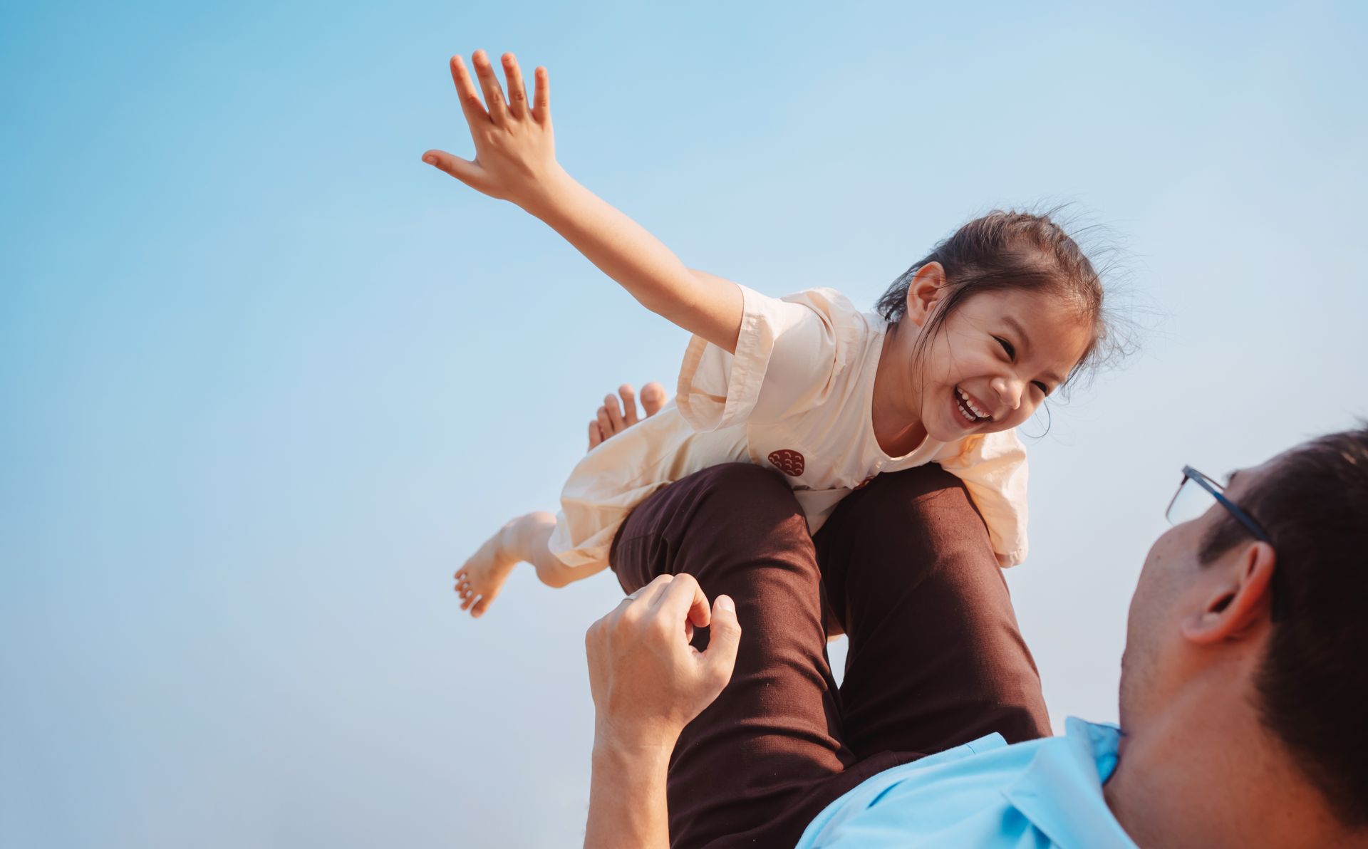 Man holds up smiling child against a blue sky, child has arms outstretched.