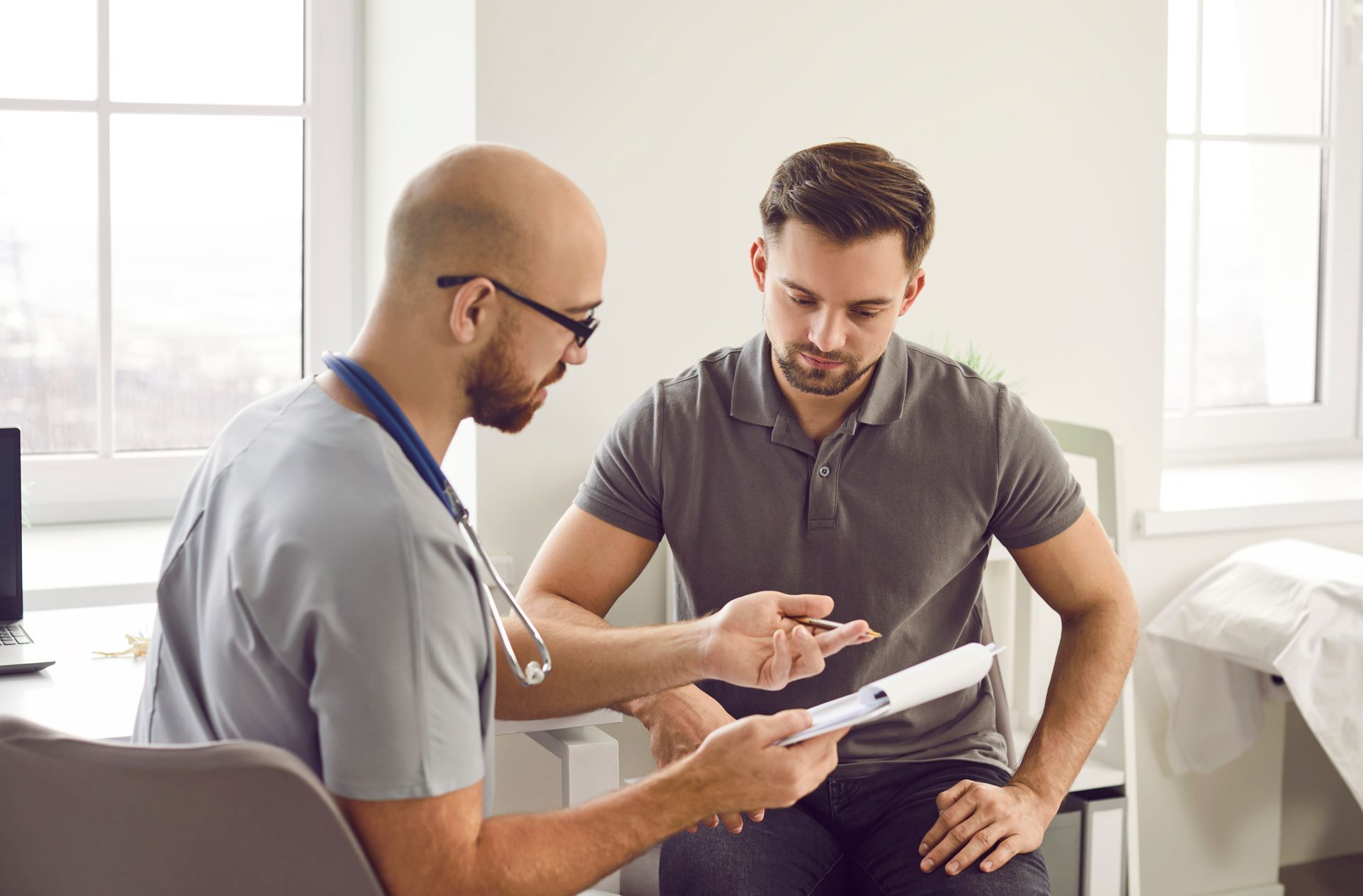 A clinician with a stethoscope discusses a medical document with a patient in a bright, modern examination room.