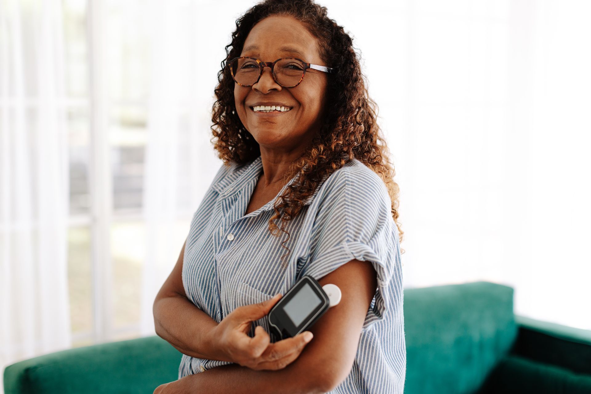 Woman smiling, holding a blood glucose meter on her arm. She is in a room with a green sofa.