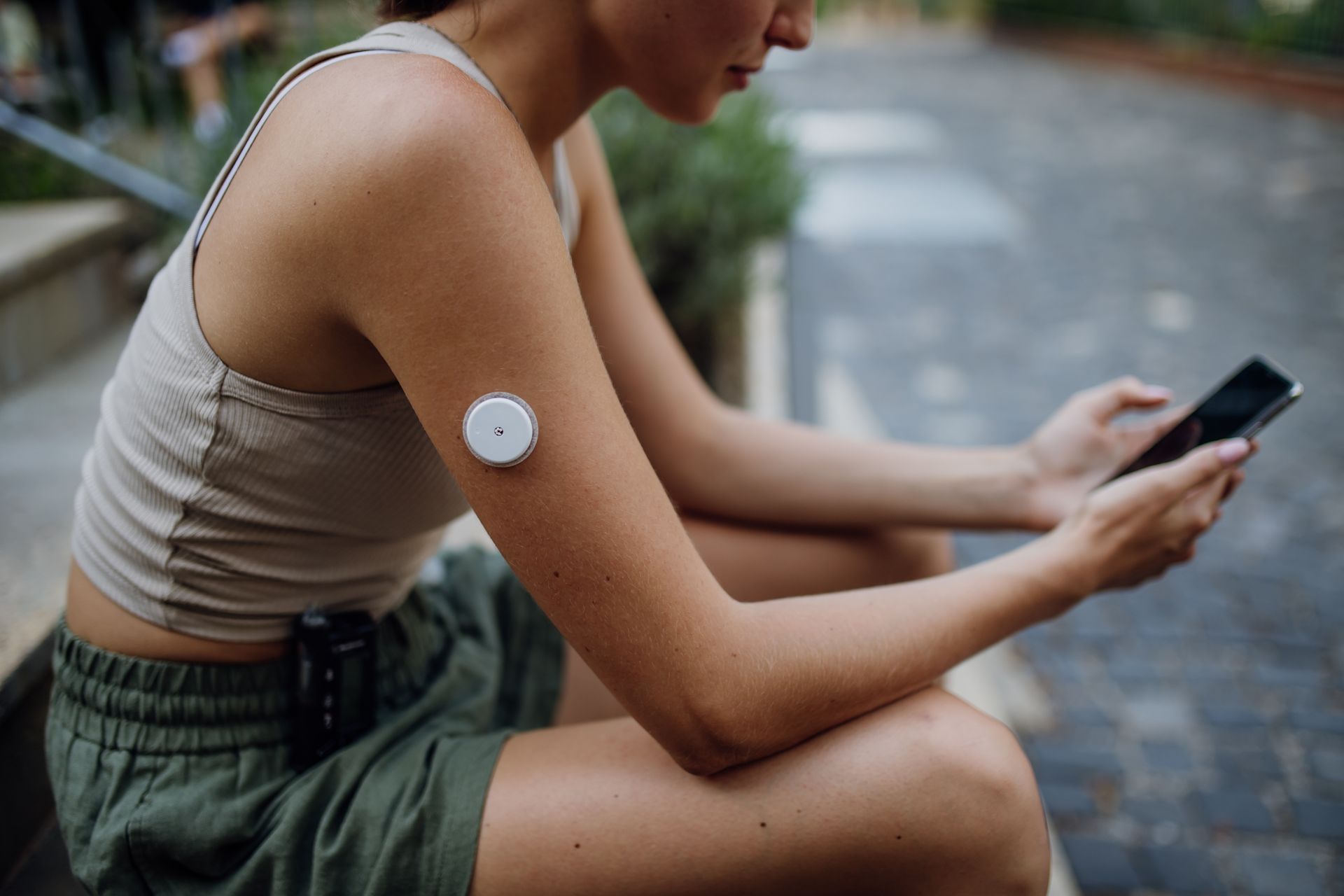Woman checks phone, connected to a continuous glucose monitor on her arm, sitting outdoors.
