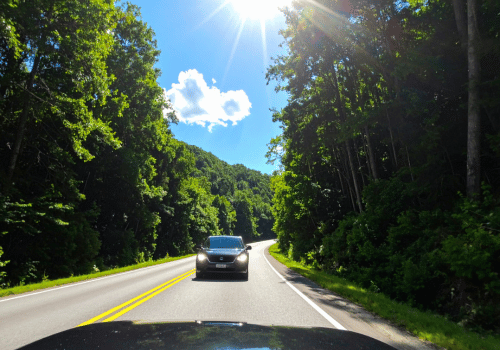 Black SUV driving on a winding forest road under bright sunlight and clear blue skies.