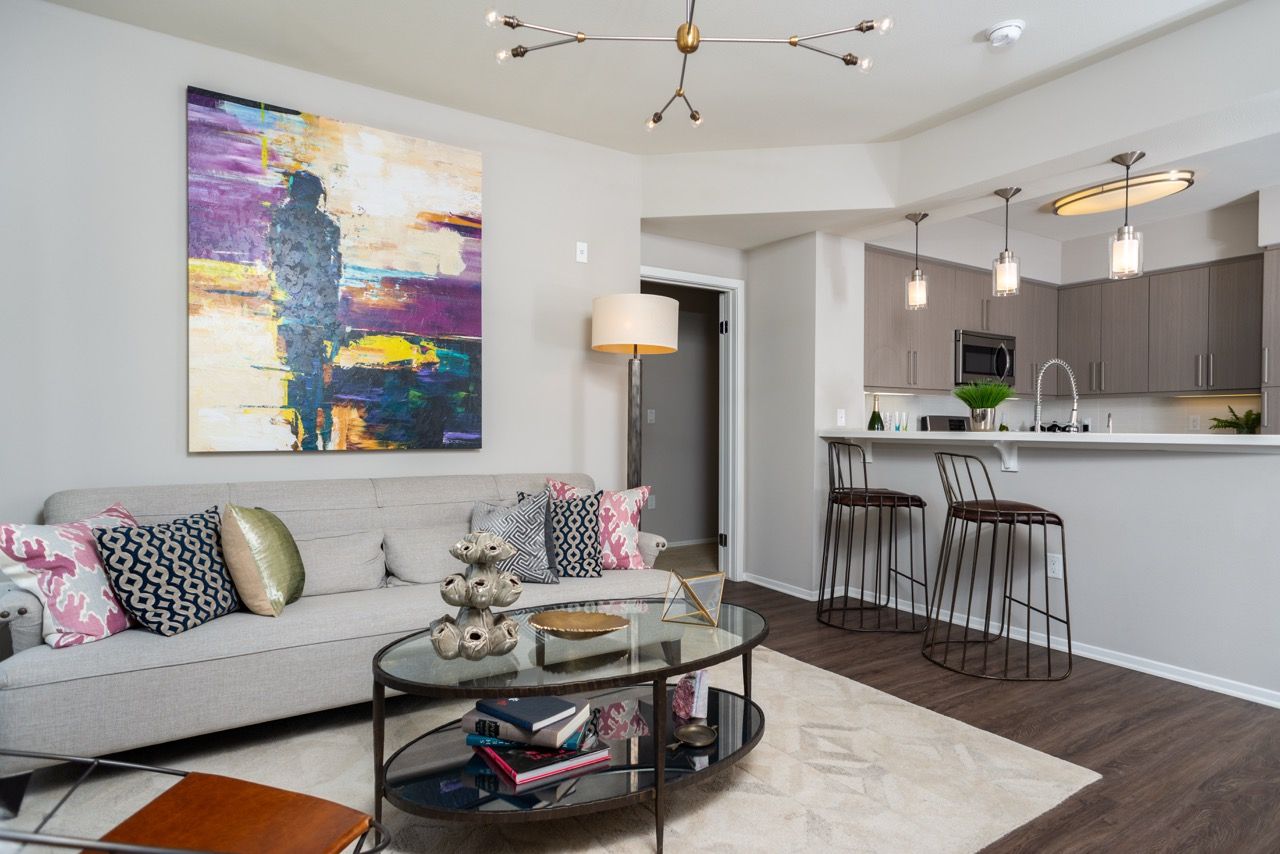 Modern apartment living room with gray sofa, colorful art, glass coffee table, and open kitchen with bar stools.