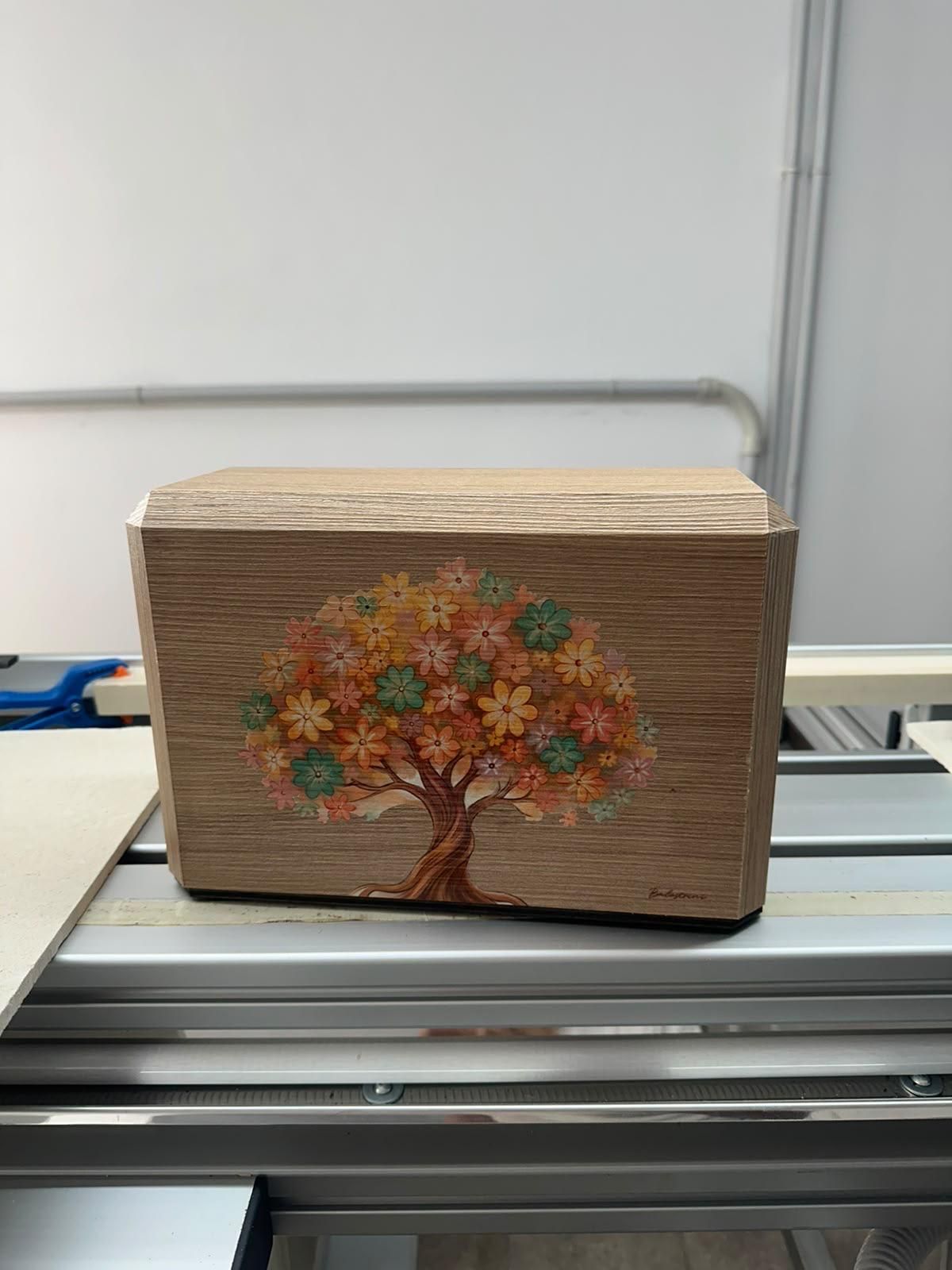 Piccola scatola di legno con un albero dipinto e fiori colorati, su un banco da lavoro davanti a una parete spoglia.