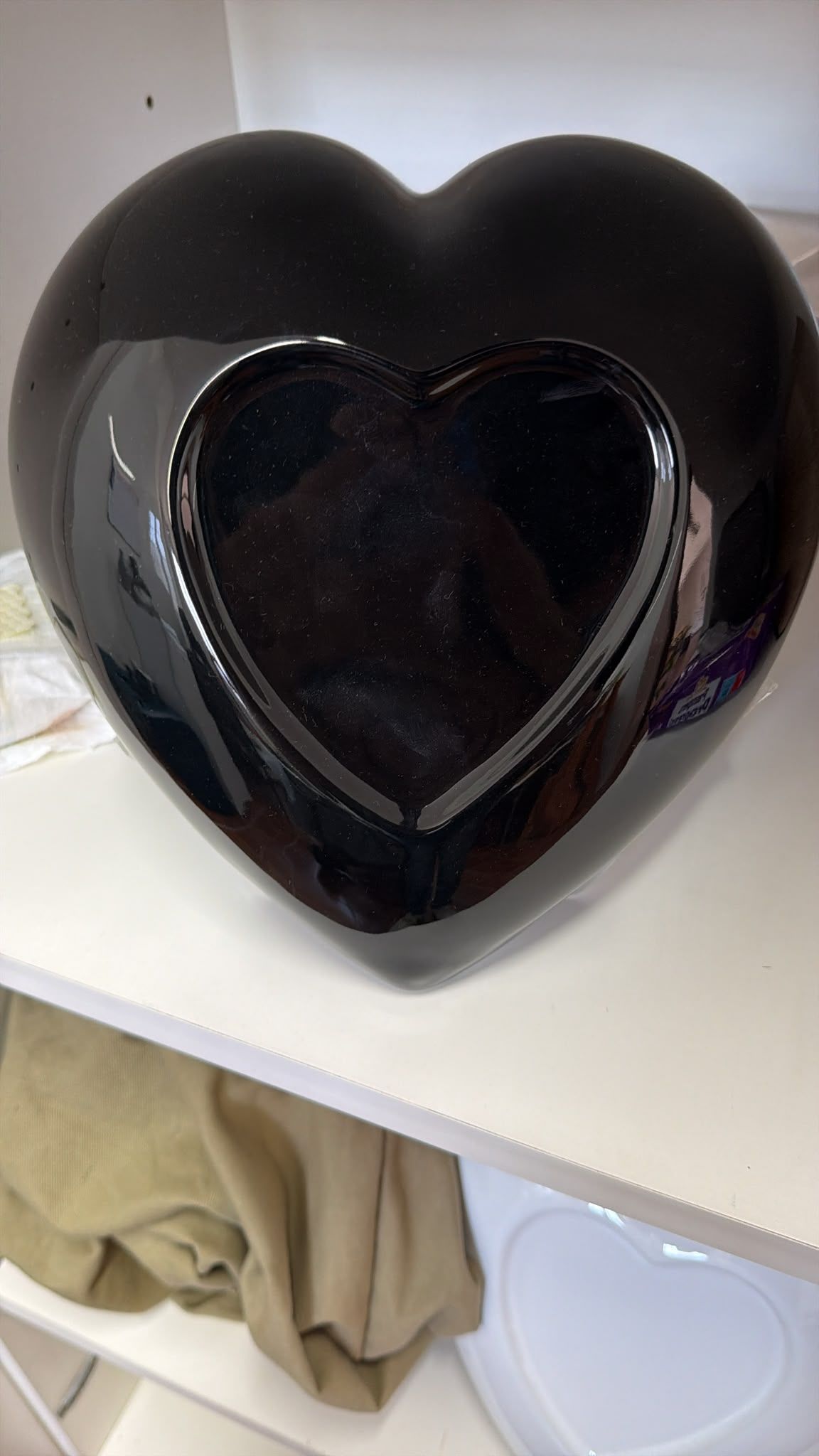 Oggetto decorativo nero a forma di cuore, con un cuoricino sul davanti, appoggiato su una mensola bianca.