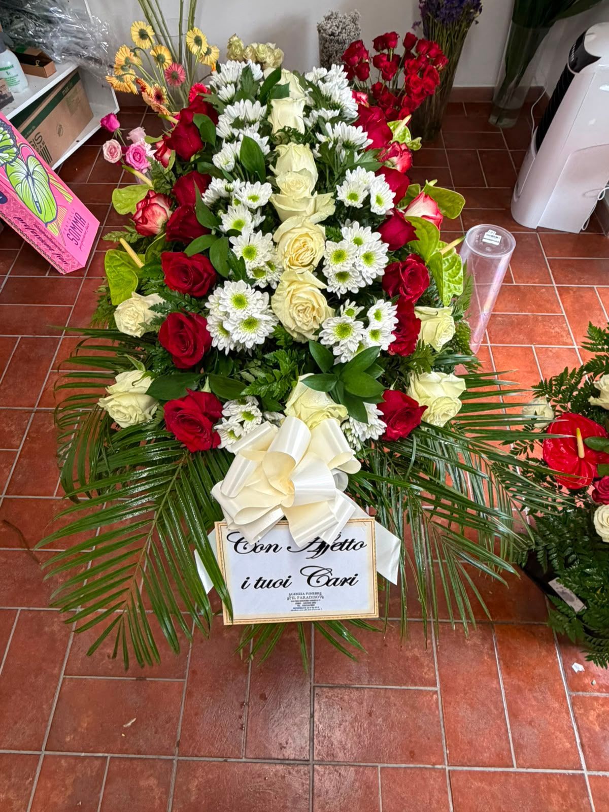 Corona funebre floreale con fiori rossi, bianchi e gialli e un nastro di condoglianze su un pavimento piastrellato