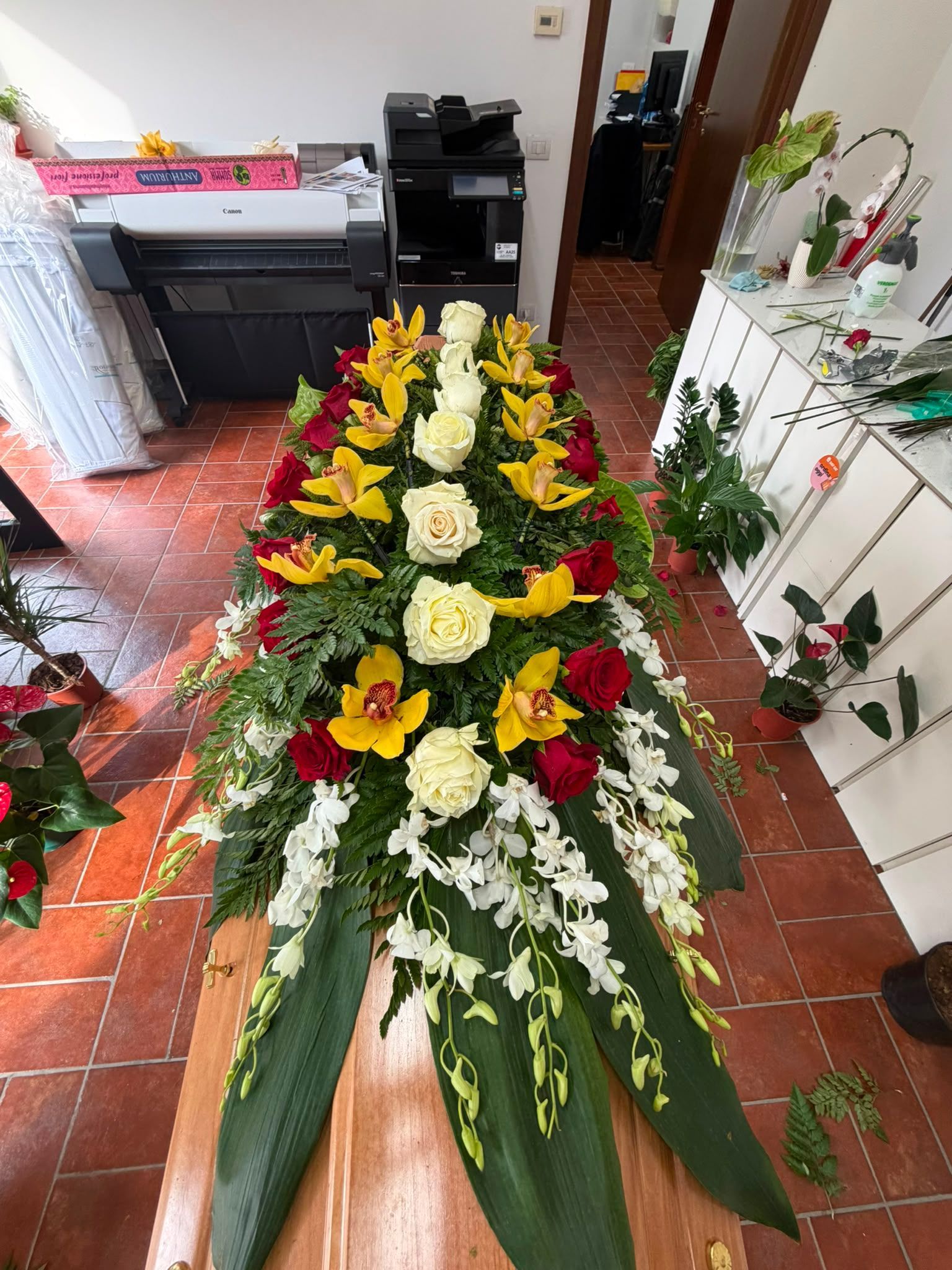 Grande corona funebre floreale con fiori gialli, bianchi e rossi, esposta al chiuso su un pavimento piastrellato.