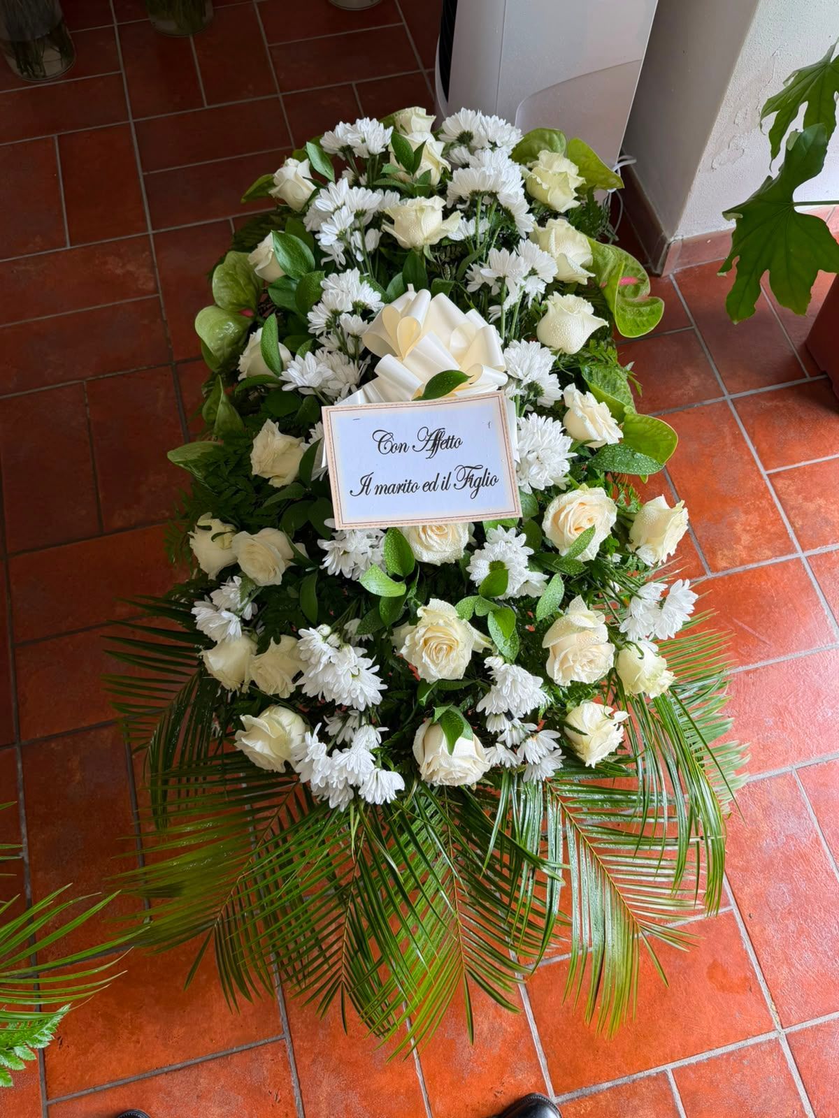 Corona di fiori bianchi con verde su un pavimento di piastrelle rosse, con un piccolo biglietto di condoglianze