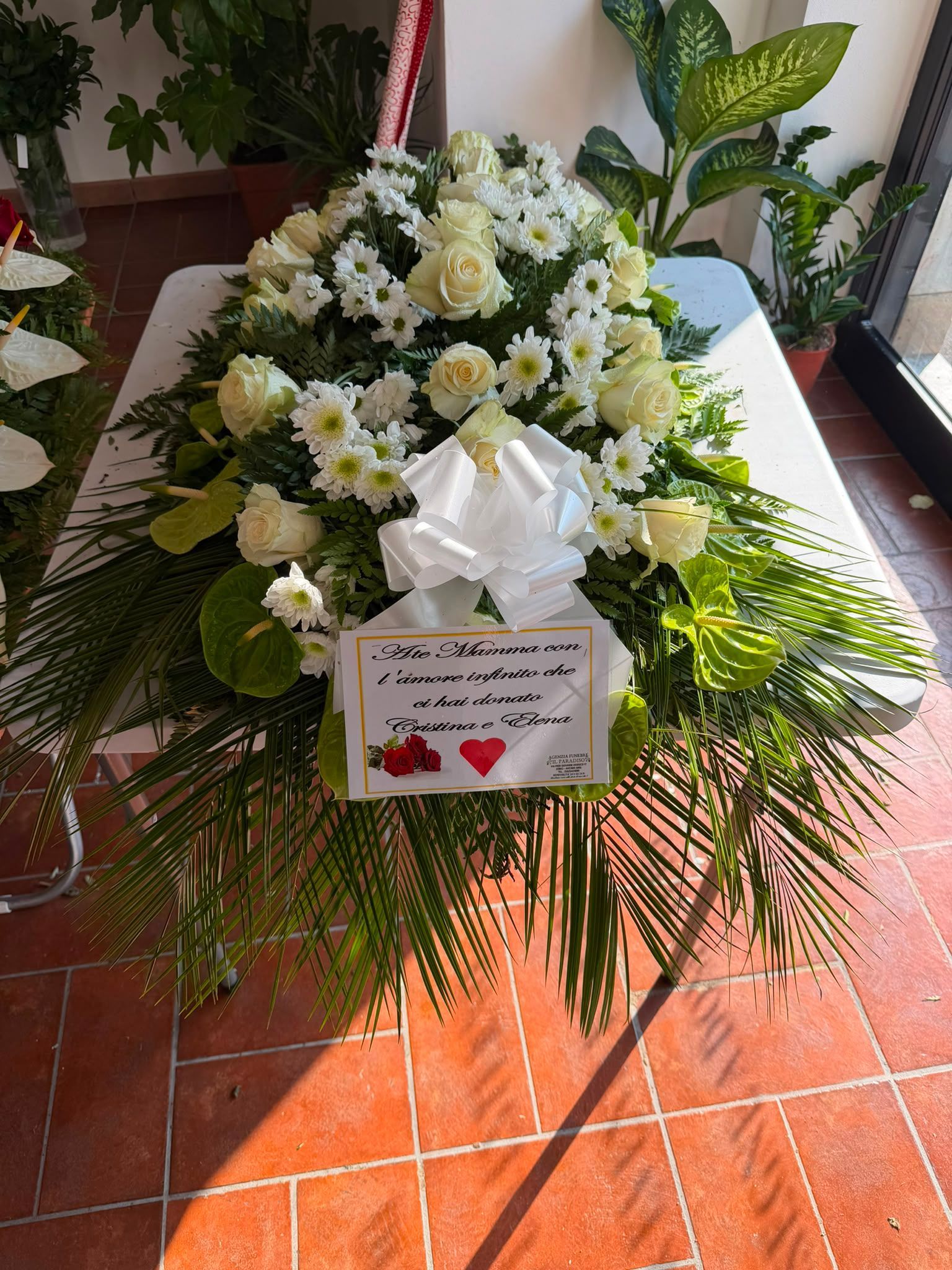 Corona funebre floreale bianca con nastro e biglietto su un portico piastrellato, circondata da foglie verdi