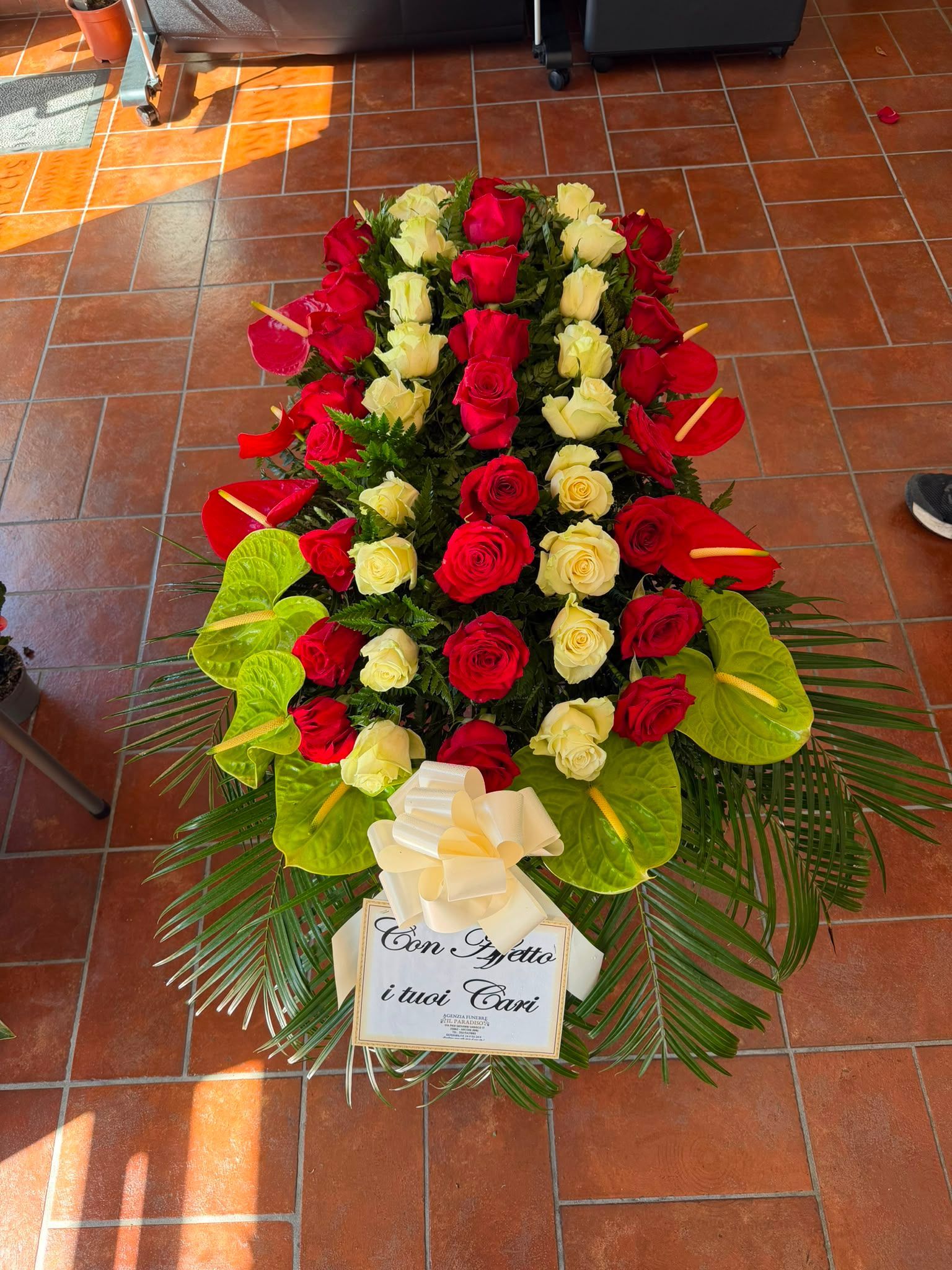 Corona funebre floreale di rose rosse, bianche e gialle con un nastro, disposta su un pavimento piastrellato.